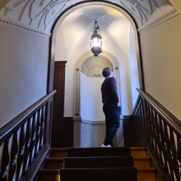 DoMpro- Remonty I Wykończenia wnętrz - Mężczyzna stojący na schodach w eleganckiej klatce schodowej z dekoracyjnym sklepieniem i oświetleniem, obserwujący ornamenty na ścianie.