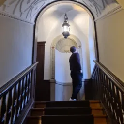 Mężczyzna stojący na schodach w eleganckiej klatce schodowej z dekoracyjnym sklepieniem i oświetleniem, obserwujący ornamenty na ścianie.