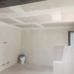 Wnętrze w trakcie prac wykończeniowych: ściany i sufit z płyt kartonowo-gipsowych, widoczne puszki elektryczne i wiadro z farbą. Etap szpachlowania.