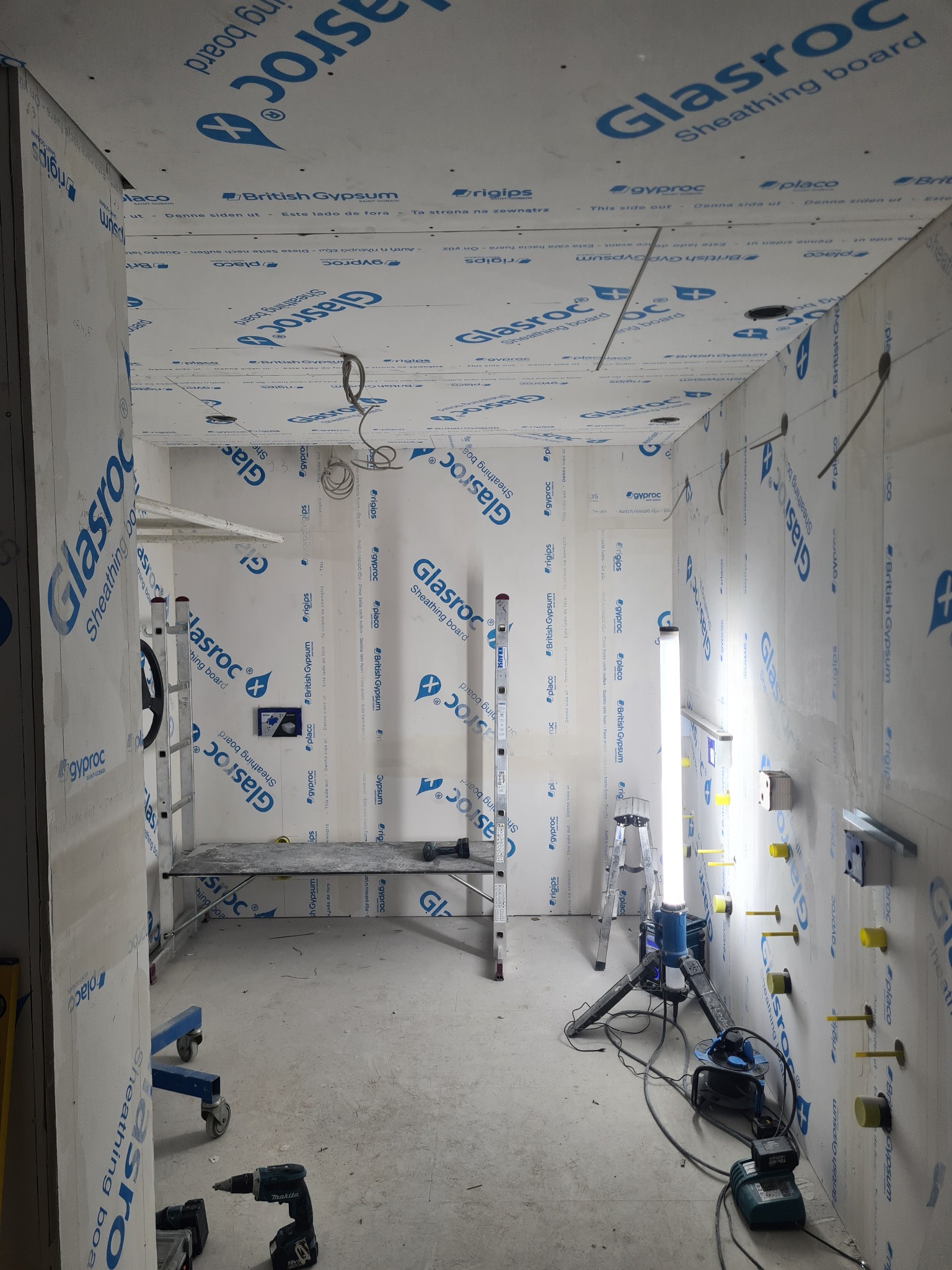Wnętrze w trakcie wykańczania, ściany obłożone płytami Glasroc, widoczne narzędzia: wiertarka, drabina, rusztowanie i lampa budowlana.