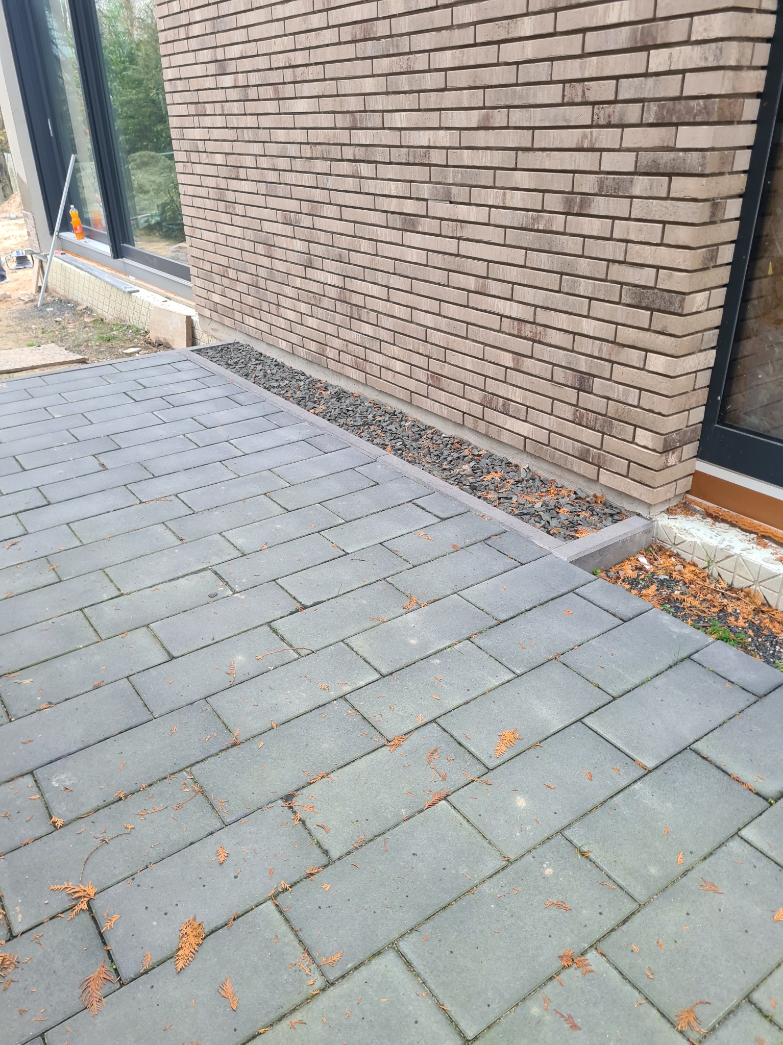 Nowoczesna elewacja budynku z cegły klinkierowej z szarym brukiem i kamiennym drenażem przy fundamencie, widoczne okno i fragment otoczenia.