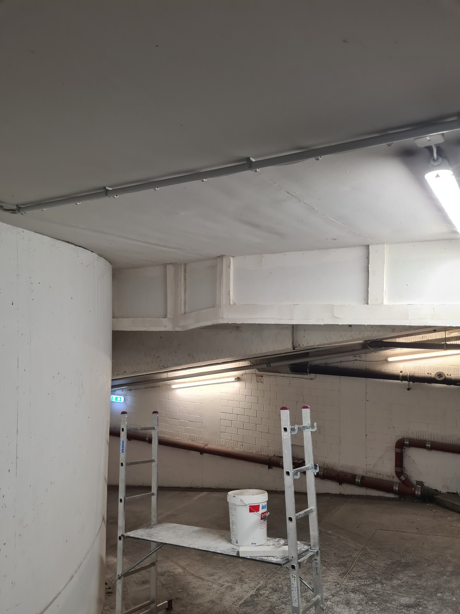 Wnętrze w trakcie malowania: biała farba na suficie i ścianach, drabina z platformą, wiadro z farbą, widoczne rury pod sufitem, surowe oświetlenie, betonowa ściana.