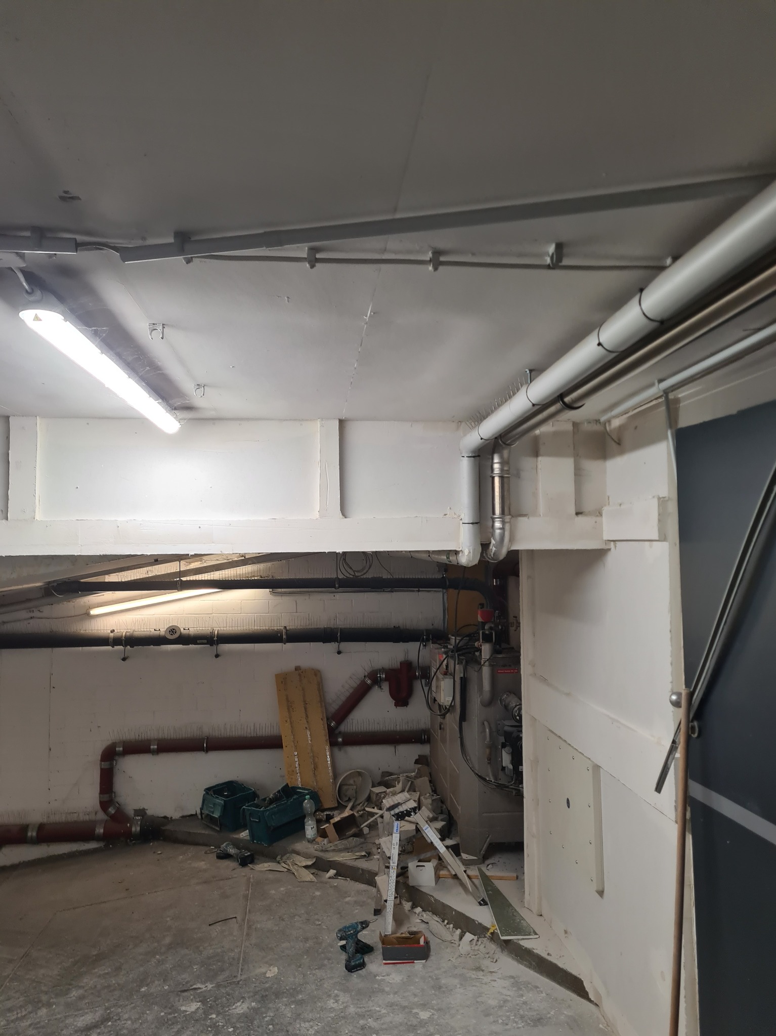 Wnętrze w trakcie remontu: widoczne rury, instalacje, narzędzia i resztki materiałów budowlanych na podłodze. Ściany częściowo wykończone, sufit z widocznymi instalacjami.