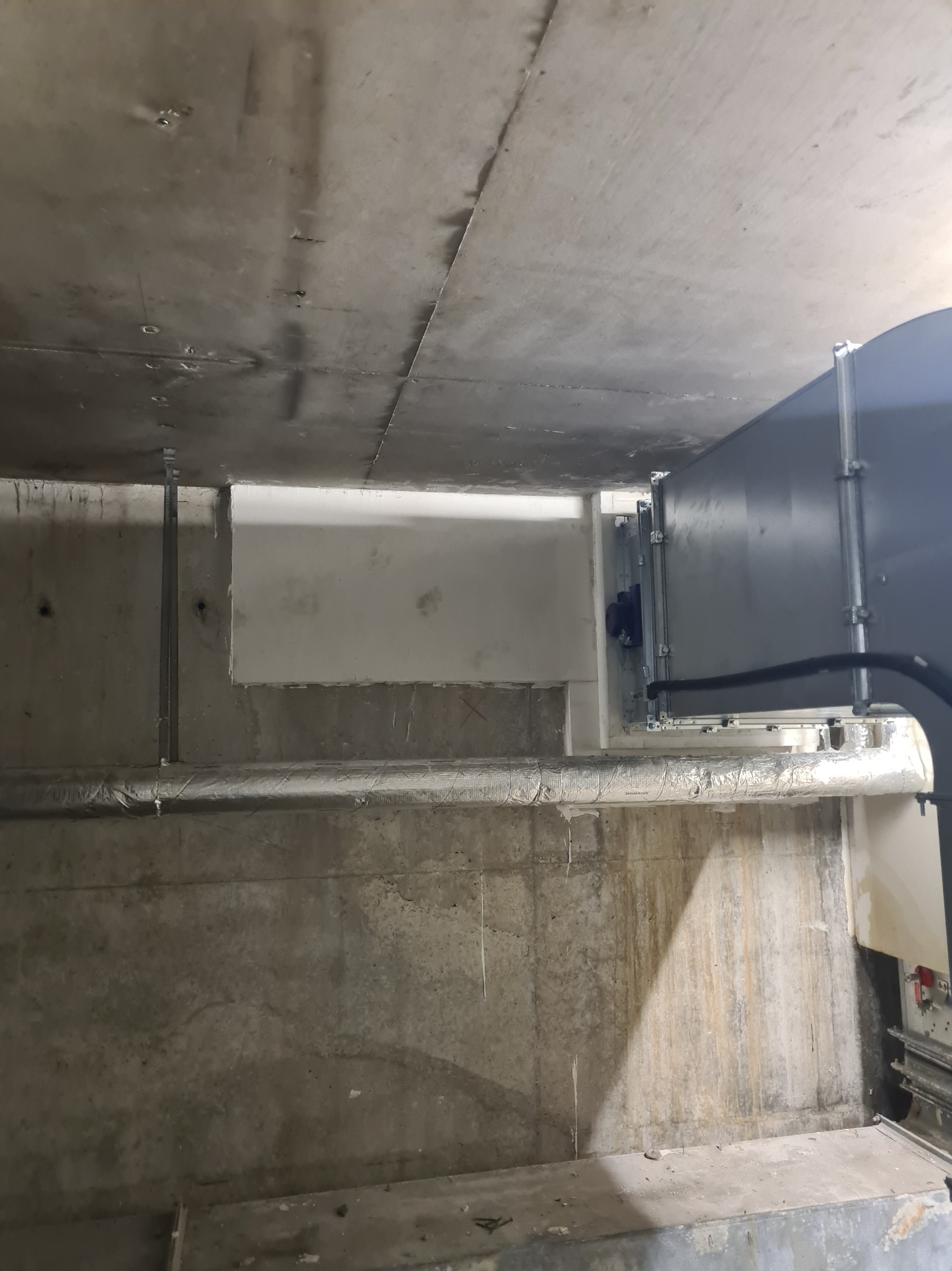 Surowy betonowy sufit z widocznymi elementami instalacji wentylacyjnej, fragment ściany z częściowo położoną gładzią i metalowe wsporniki.