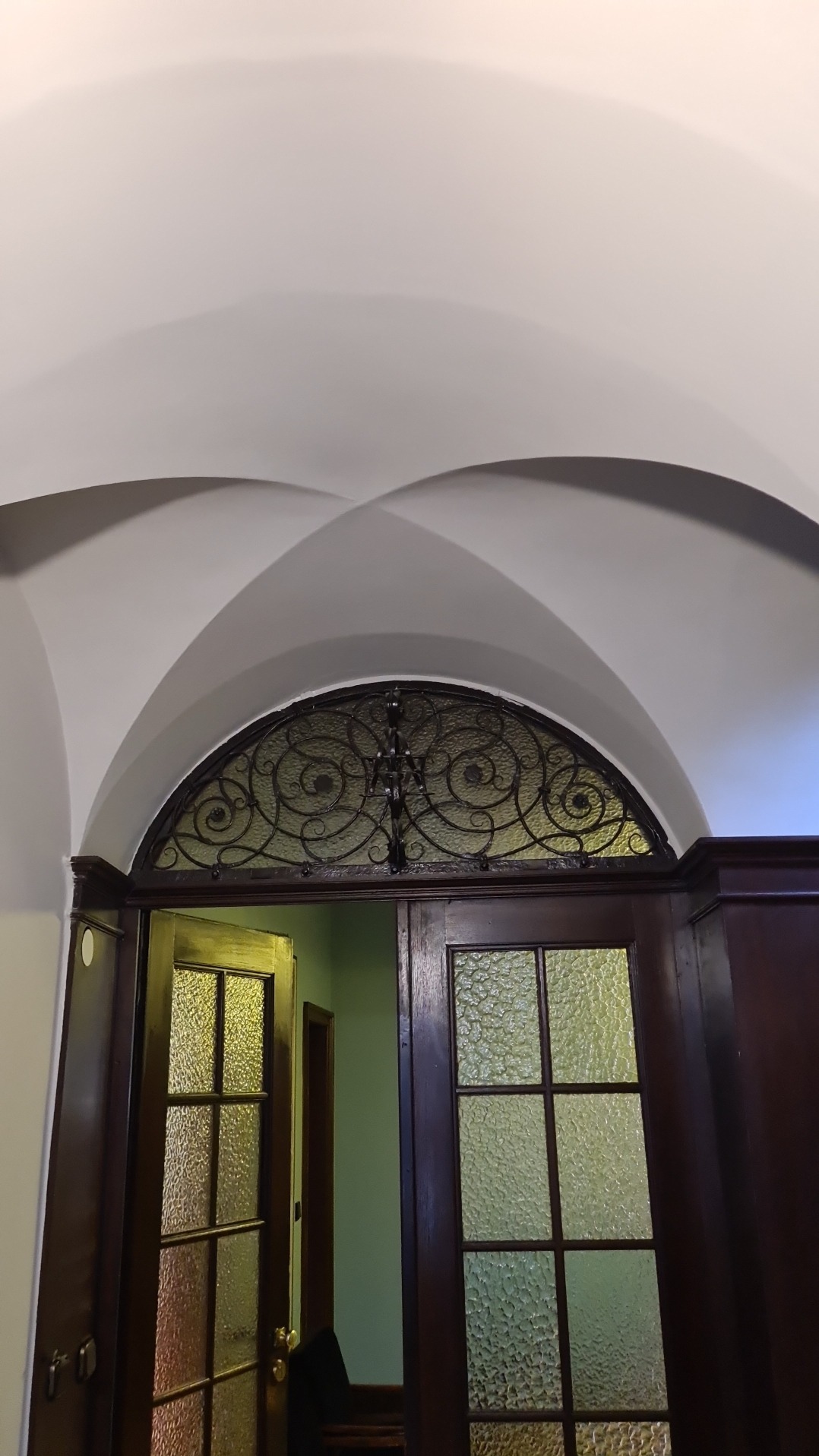 Drewniane drzwi z ozdobną, metalową kratą w górnej części, widoczne pod łukowatym sufitem.