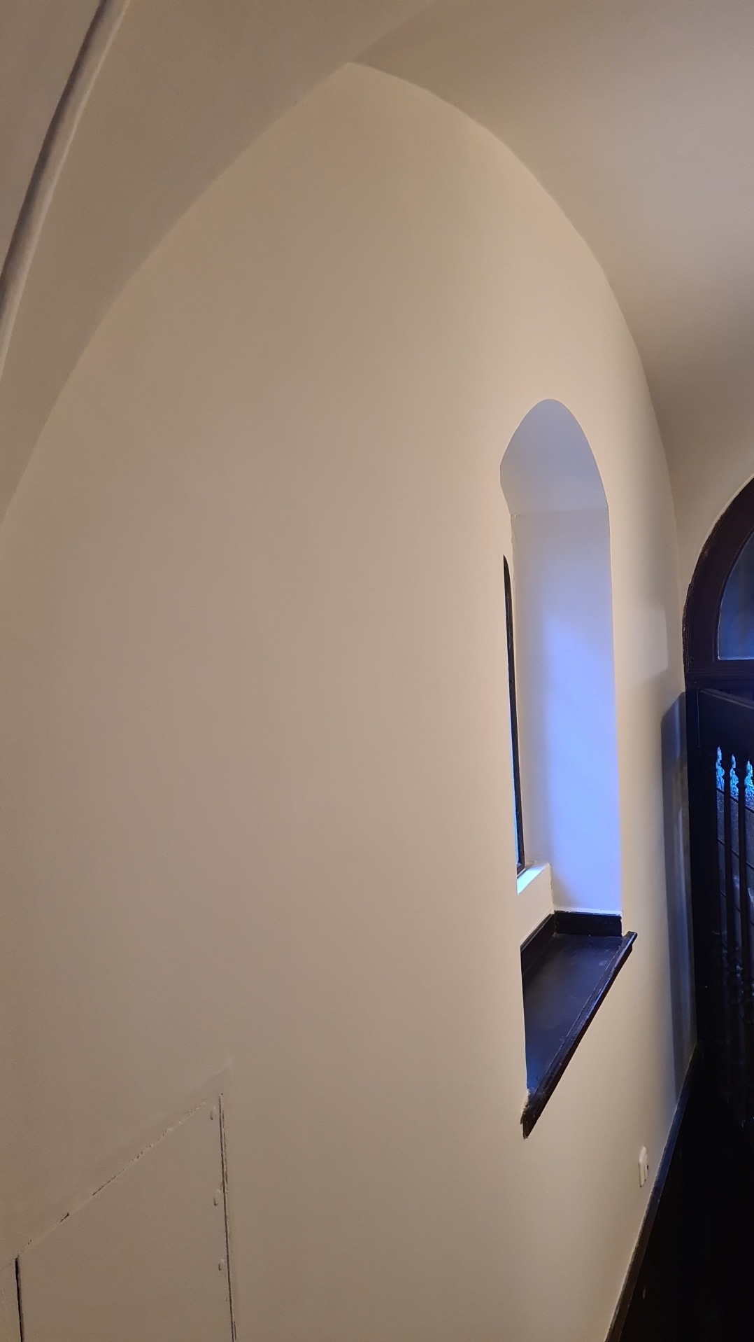Wnętrze z łukowatym oknem i świeżo pomalowaną, gładką ścianą w odcieniach bieli, widoczne detale wykończeniowe i architektoniczne.