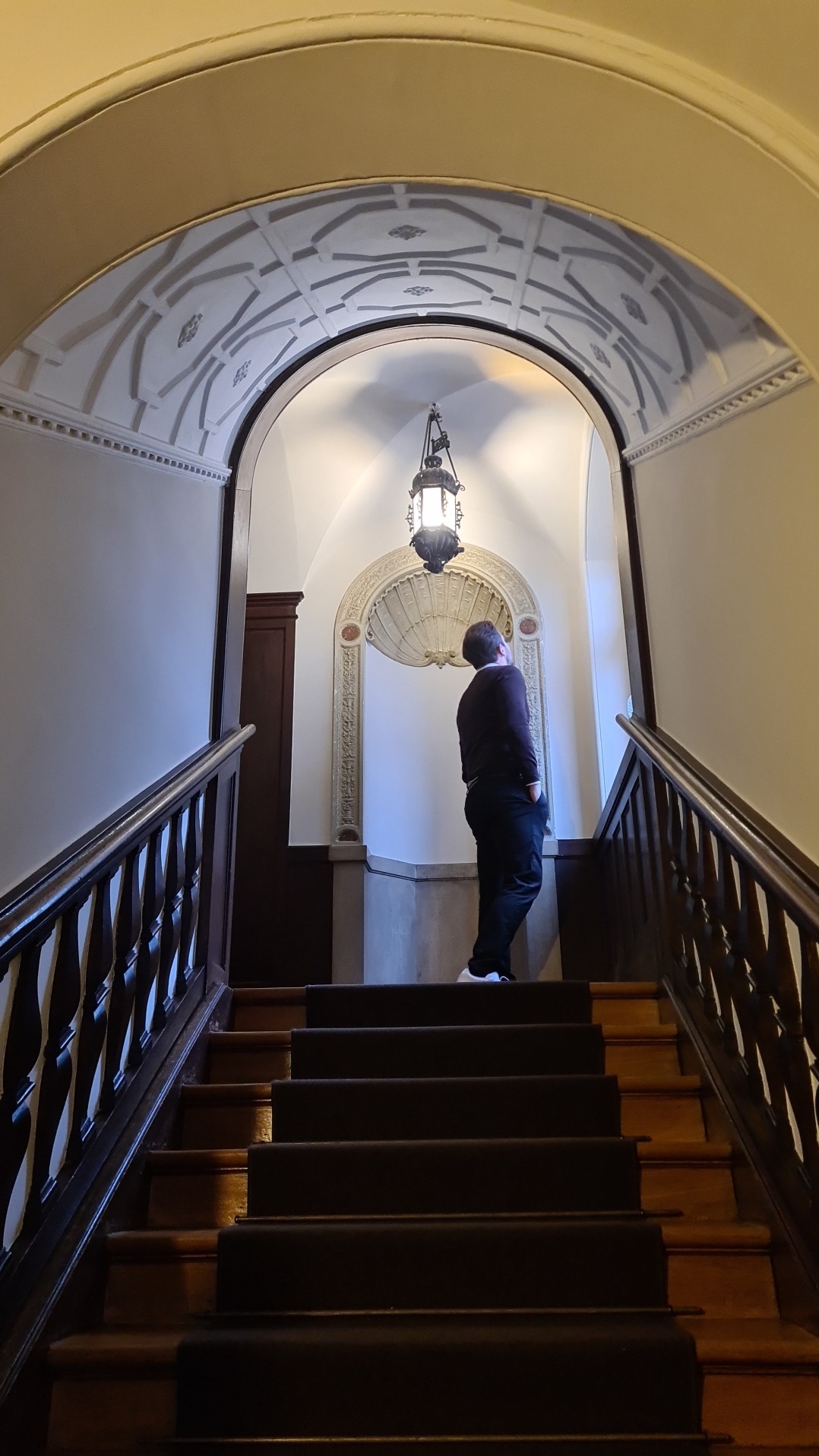Mężczyzna stojący na schodach w eleganckiej klatce schodowej z dekoracyjnym sklepieniem i oświetleniem, obserwujący ornamenty na ścianie.