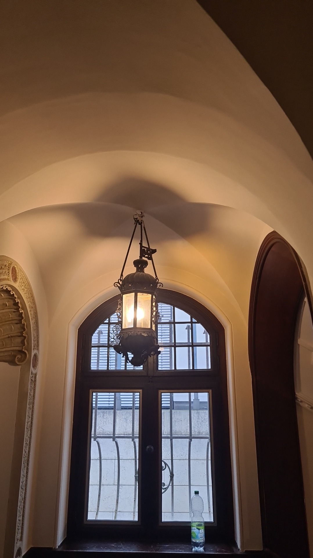 Wnętrze z wysokim sklepieniem łukowym, okno z dekoracyjną kratą i wisząca, stylizowana lampa; na parapecie stoi butelka wody.