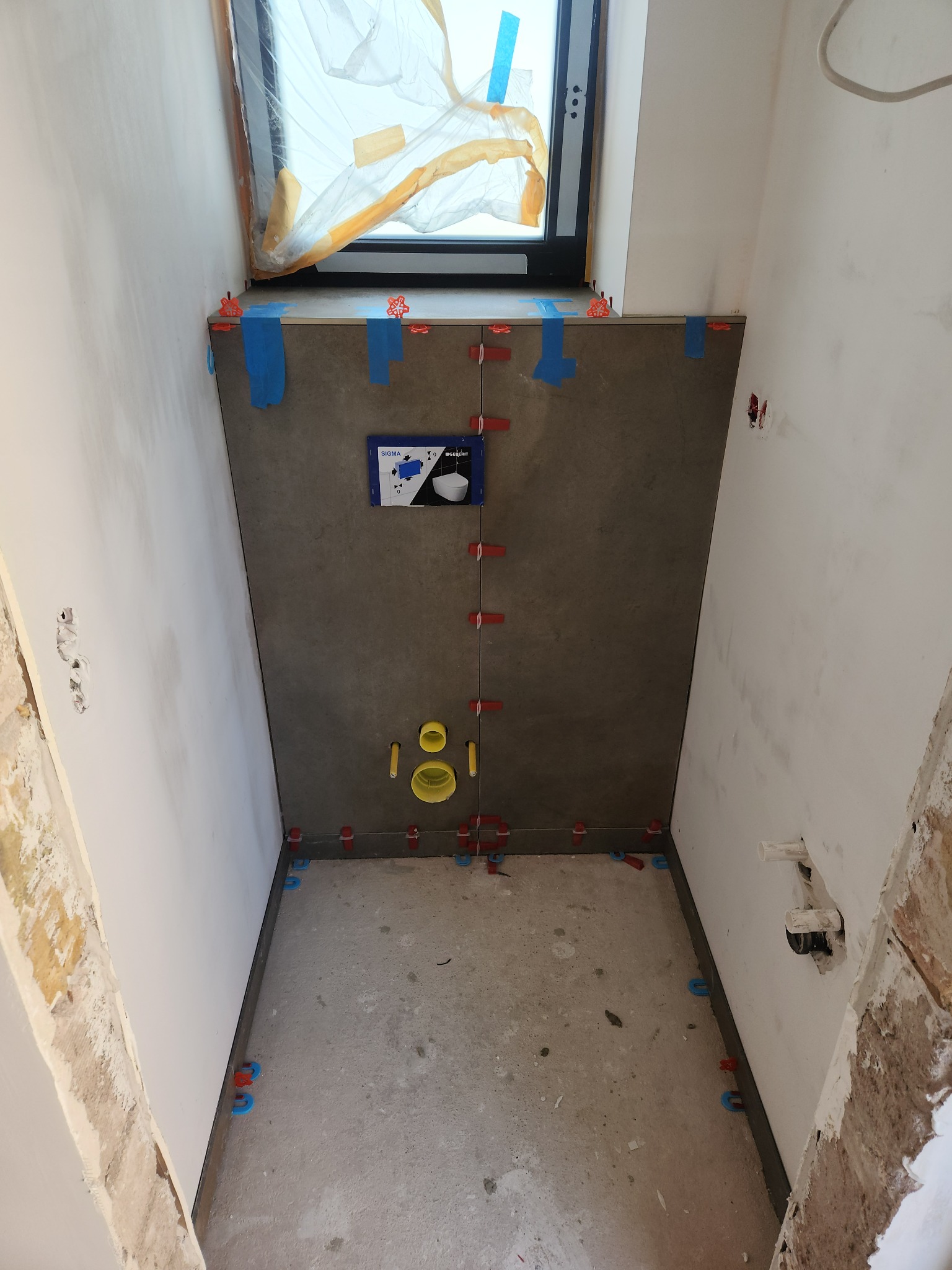 Montaż ciemnych, matowych płytek w małej łazience z oknem. Widoczne elementy instalacji podtynkowej i akcesoria do poziomowania płytek. Etap prac przed fugowaniem.