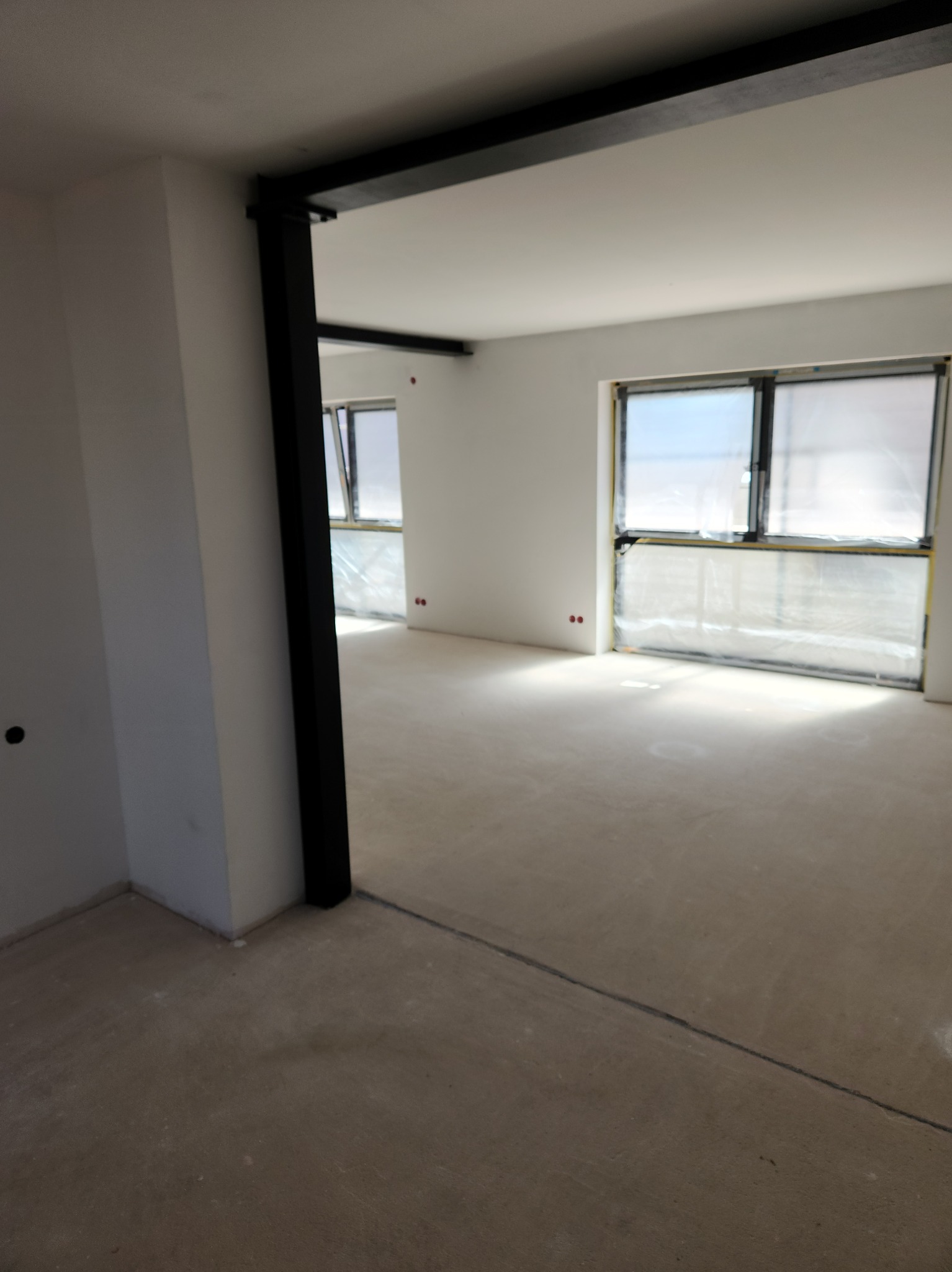 Wnętrze w trakcie prac: surowa podłoga, okna zabezpieczone folią, widoczne ślady po gładzi. Czarna rama drzwi oddziela pomieszczenia.