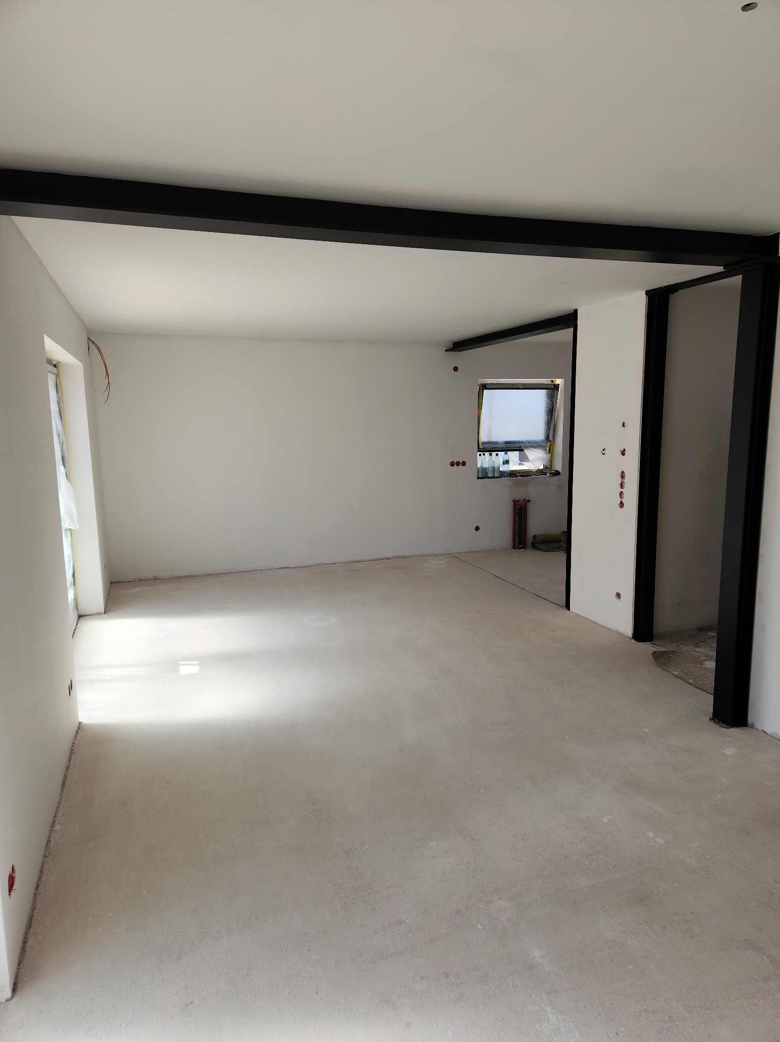 Surowe wnętrze z białymi ścianami, betonową podłogą i czarnymi elementami konstrukcyjnymi. Widoczne przygotowania do dalszych prac wykończeniowych.