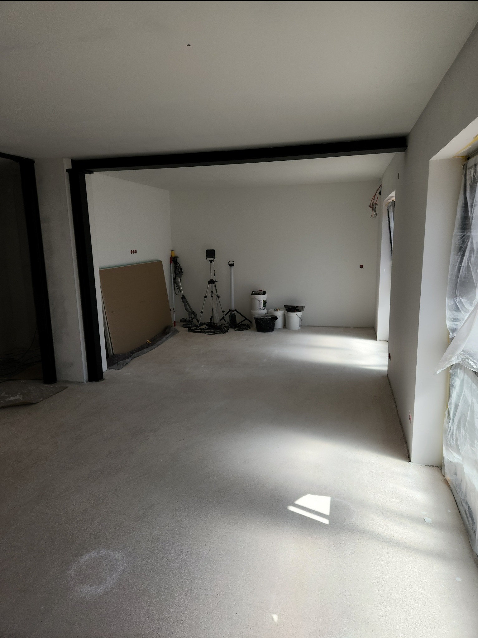 Wnętrze w trakcie prac wykończeniowych: świeżo wylana posadzka, przygotowane płyty gips-karton, widoczne ślady po pomiarach na podłodze, sprzęt malarski w tle.