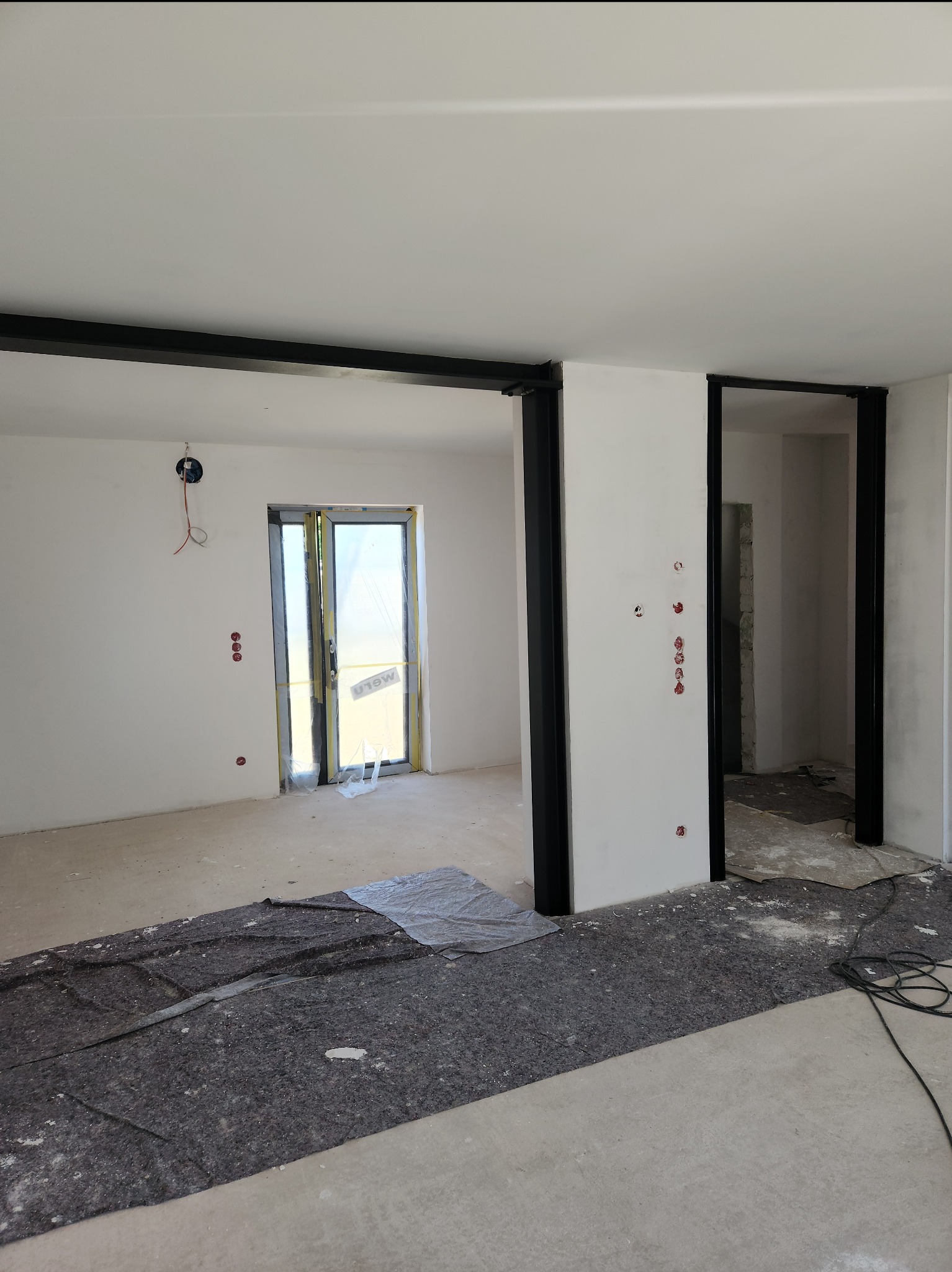 Wnętrze w trakcie prac wykończeniowych: ściany przygotowane do malowania, widoczne instalacje elektryczne i zabezpieczone drzwi balkonowe.