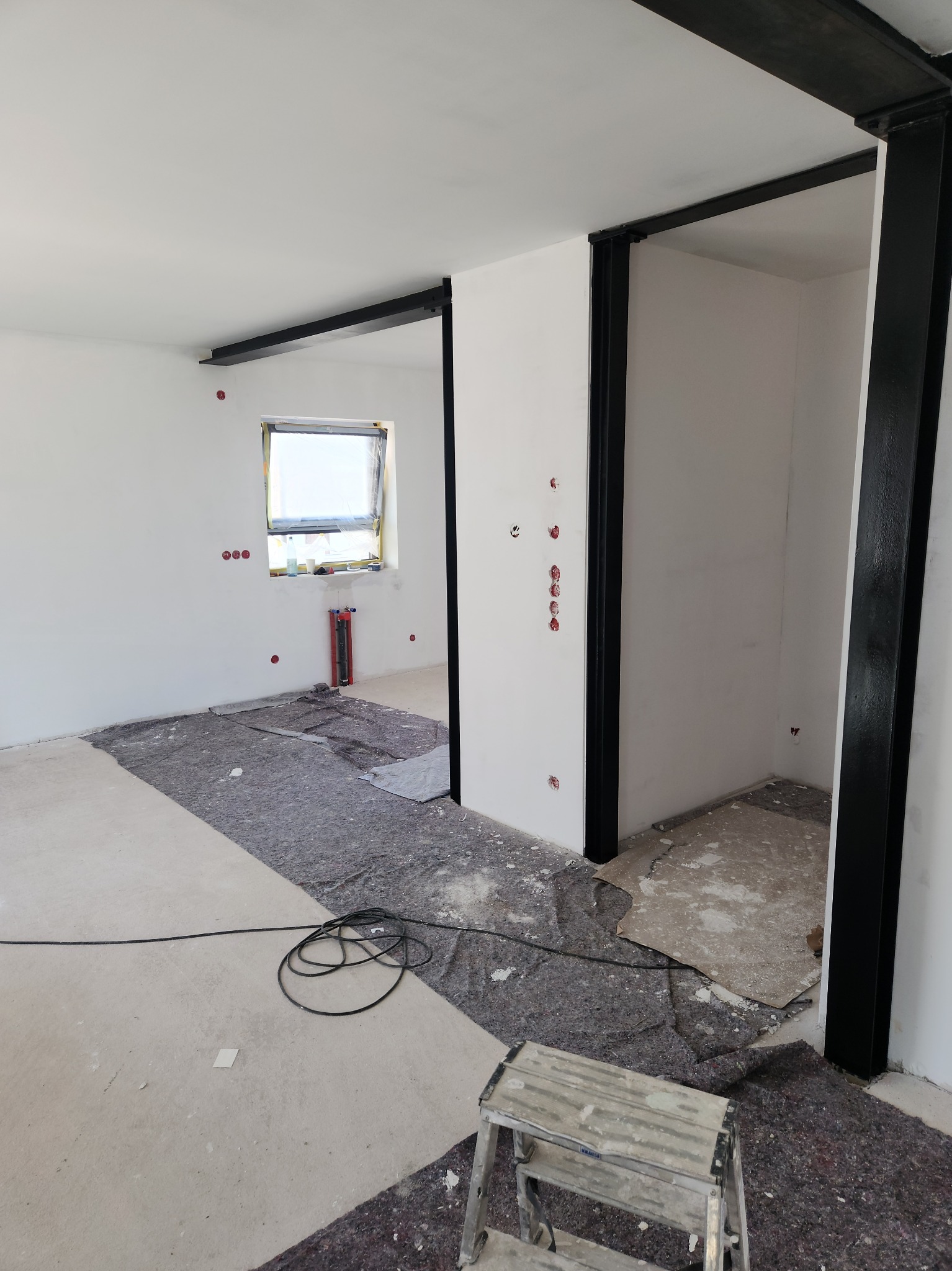 Wnętrze w trakcie remontu: białe ściany, czarne metalowe ramy, zabezpieczona podłoga, widoczne instalacje, drabina malarska. Stan surowy, przygotowanie do dalszych prac wykończeniowych.