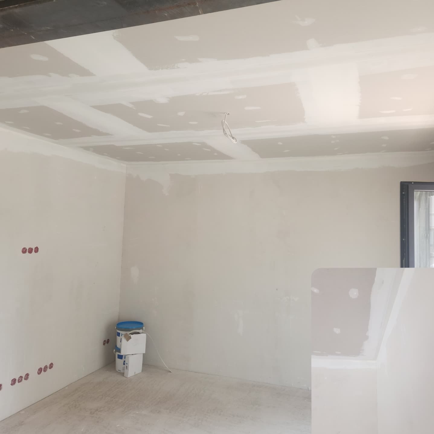 Wnętrze w trakcie prac wykończeniowych: ściany i sufit z płyt kartonowo-gipsowych, widoczne puszki elektryczne i wiadro z farbą. Etap szpachlowania.