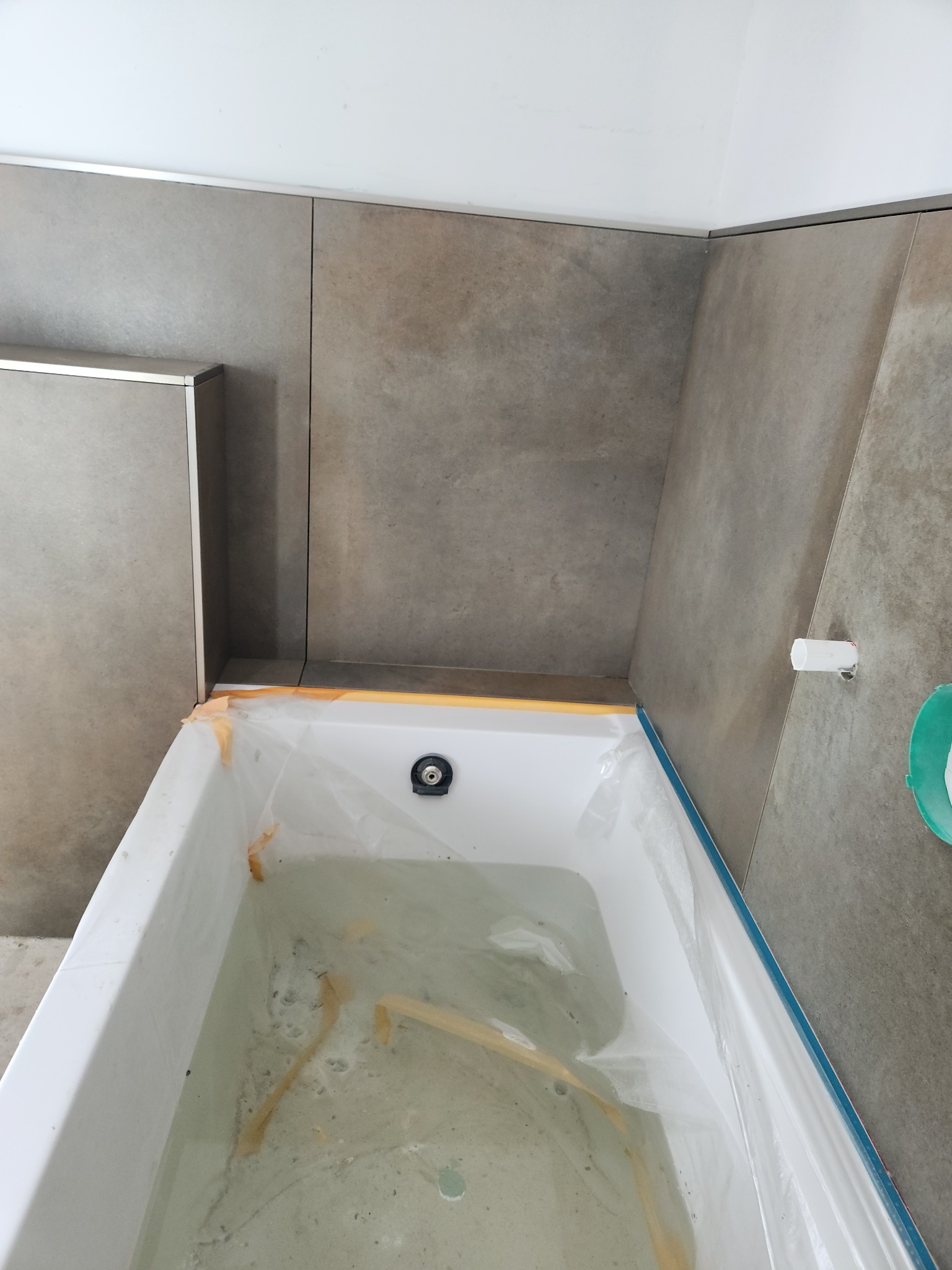 Wykończona łazienka z nową wanną zabezpieczoną folią, szare płytki na ścianach z metalowymi listwami, widok z góry.