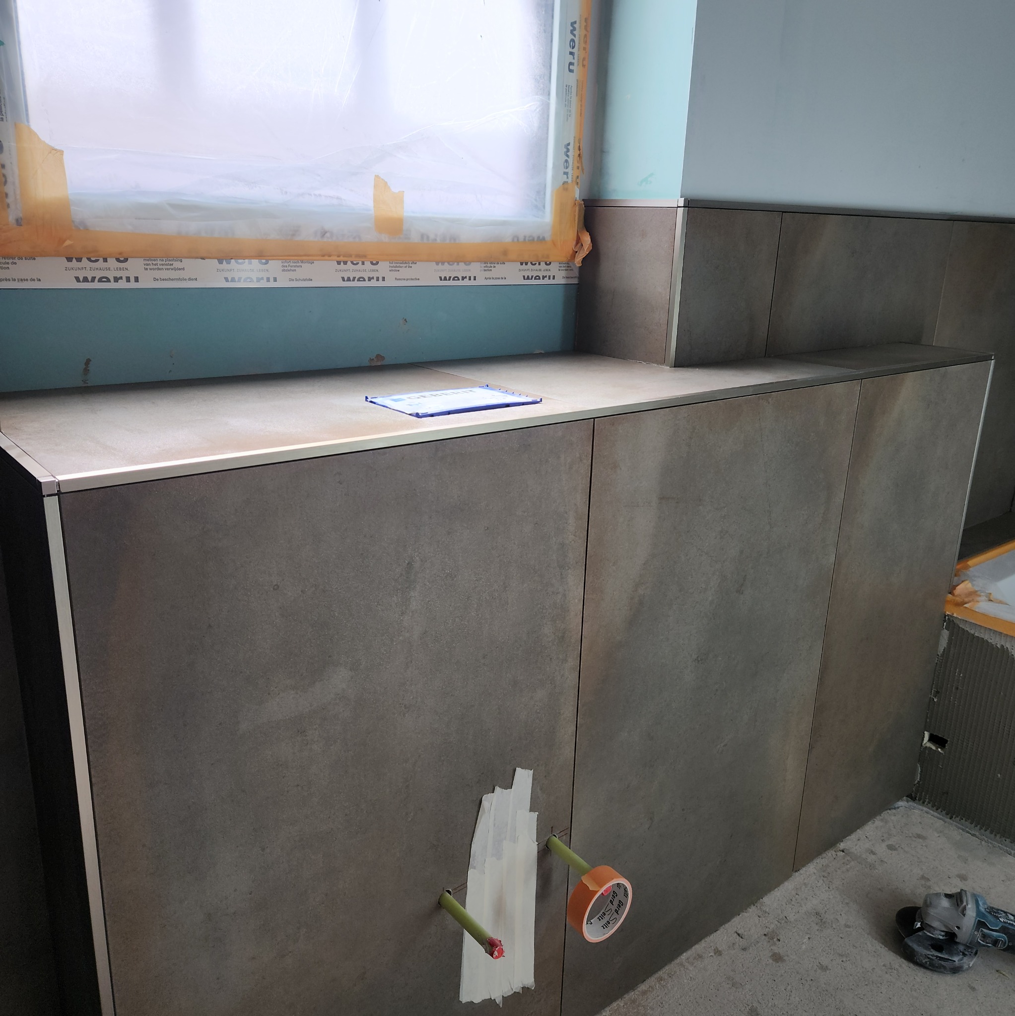 Szara zabudowa z płytek w łazience w trakcie montażu, widoczne rury i narzędzia. Okno zabezpieczone folią ochronną, na blacie leży dokumentacja techniczna.