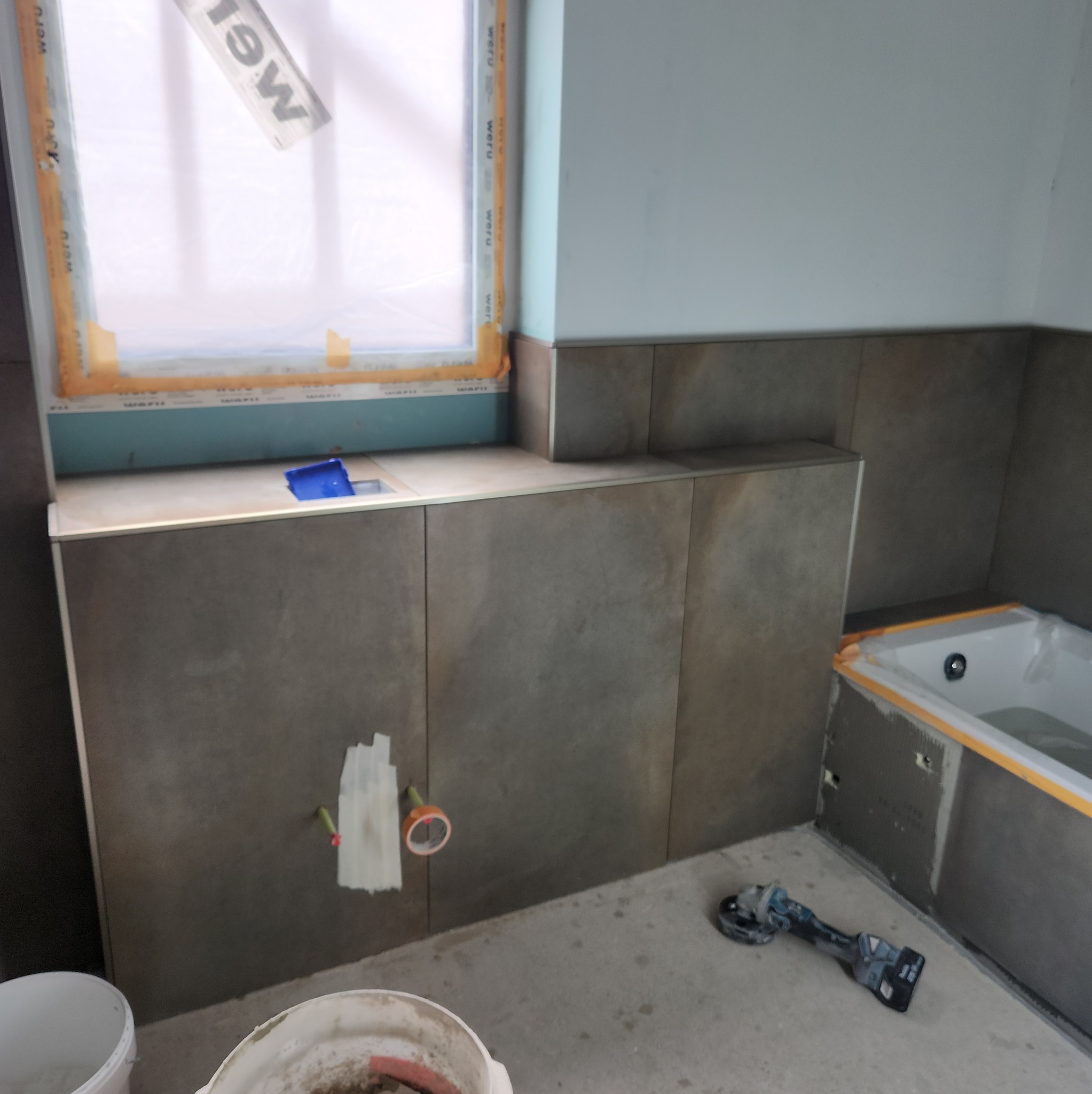 Układanie płytek w łazience, widoczne narzędzia glazurnicze, okno zabezpieczone folią. Płytki w odcieniach szarości, wanna w tle. Prace w toku, widok na fragment ściany.