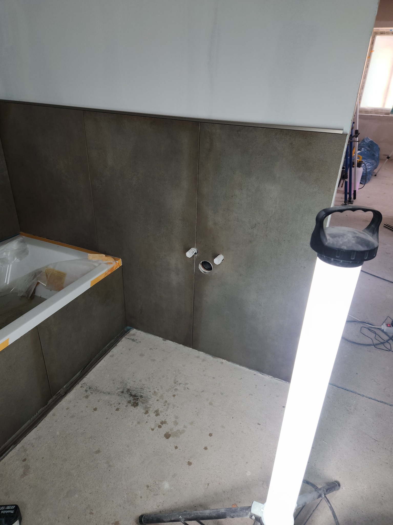 Montaż ciemnych płytek w łazience, widoczne otwory na armaturę, zabezpieczona wanna i lampa budowlana. Prace glazurnicze w trakcie realizacji.