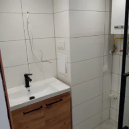 Nowoczesna łazienka z białymi płytkami, drewnianą szafką pod umywalką i czarną armaturą. Widoczne przewody elektryczne i rury gazowe.