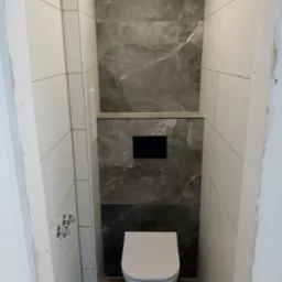 Nowoczesna toaleta z zabudowaną spłuczką, wykończona białymi i szarymi płytkami imitującymi marmur. Widoczne chromowane zawory i fragment niedokończonego wykończenia.