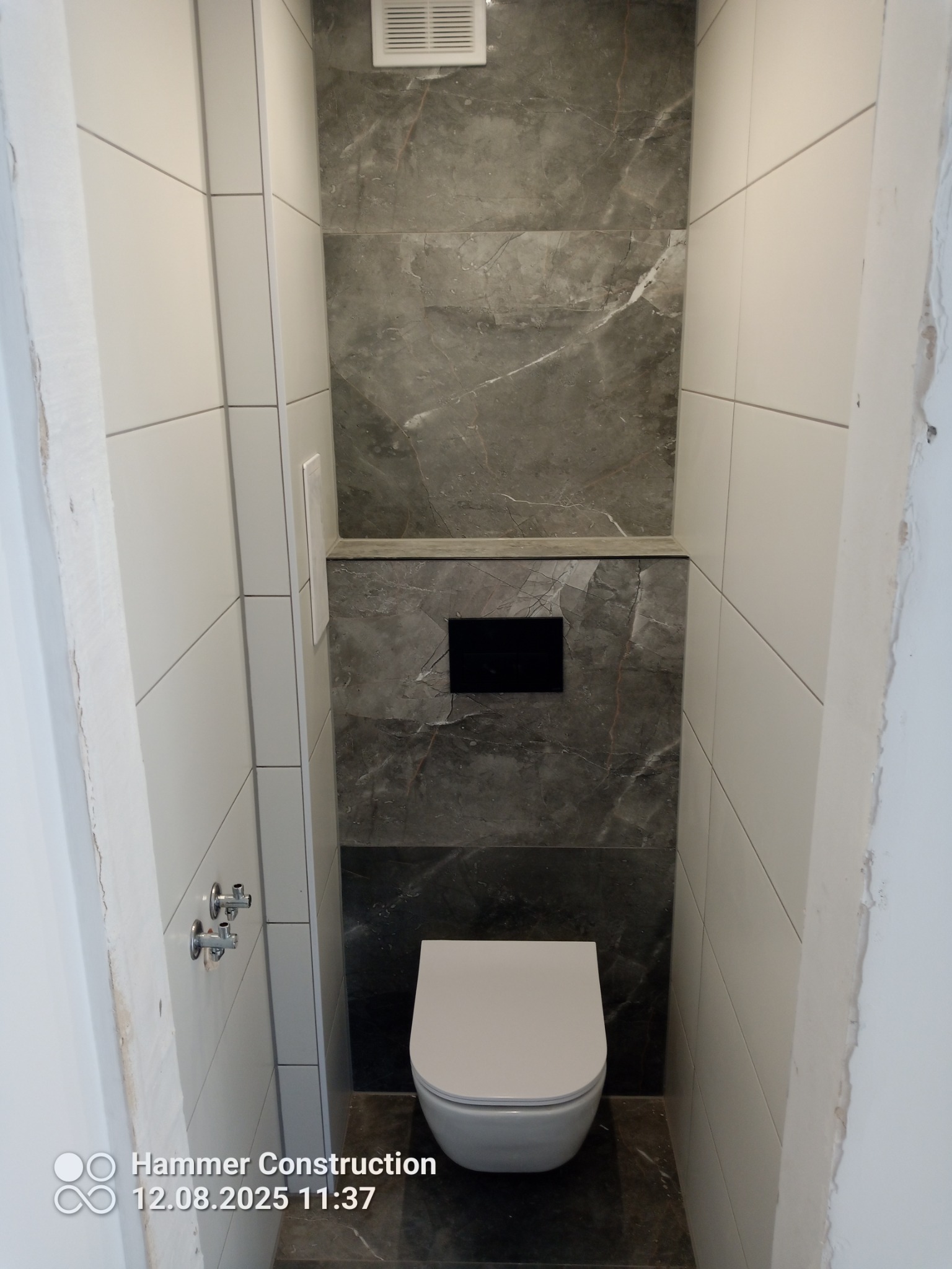 Nowoczesna toaleta z zabudowaną spłuczką, wykończona białymi i szarymi płytkami imitującymi marmur. Widoczne chromowane zawory i fragment niedokończonego wykończenia.