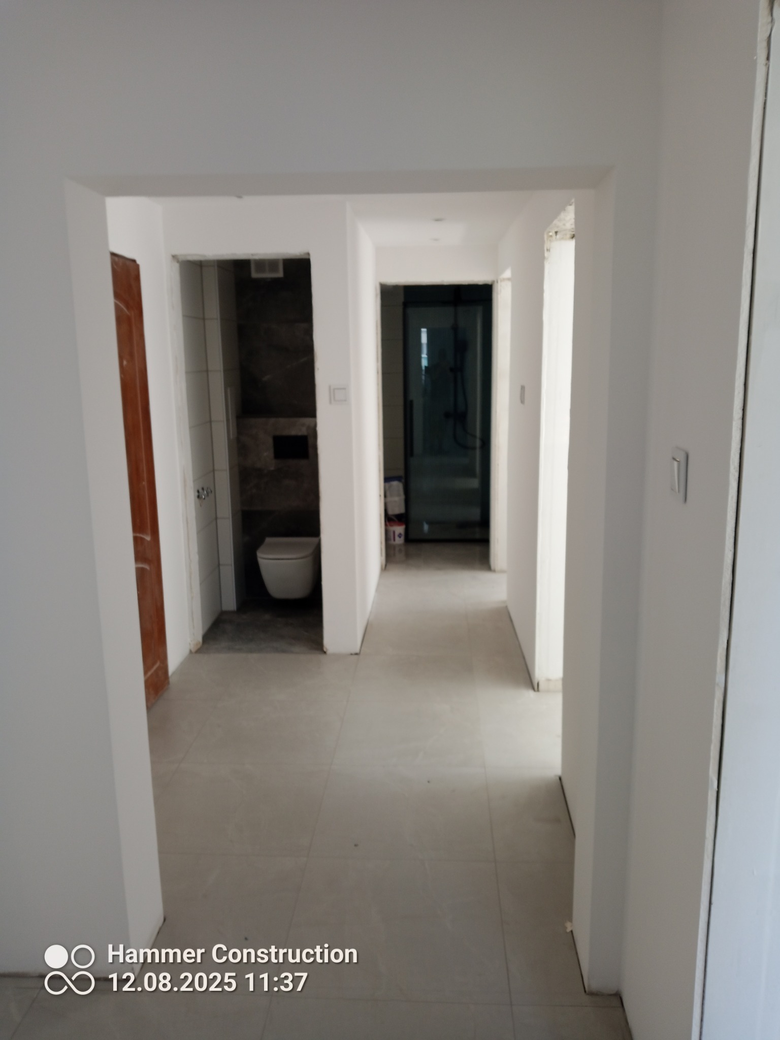 Wnętrze w trakcie wykańczania: widok na korytarz z wejściami do łazienki i pokoju z kabiną prysznicową. Szare płytki na podłodze, białe ściany, surowy stan wykończenia.