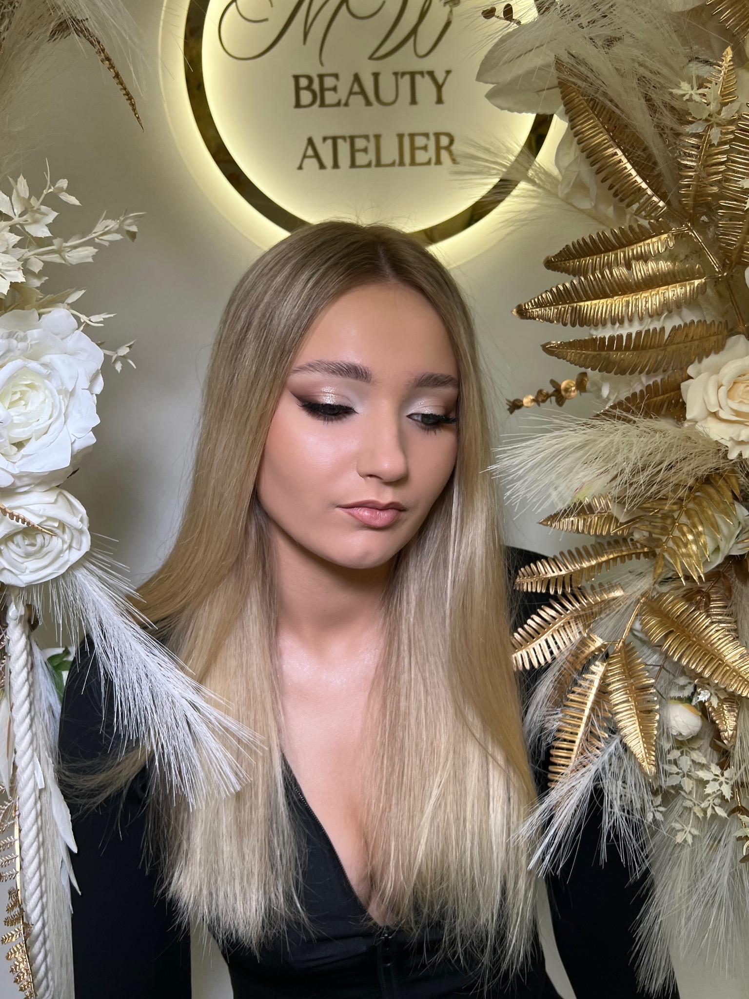 Portret blondynki z profesjonalnym, błyszczącym makijażem oczu w złotych i brązowych tonacjach, pozującej na tle dekoracji z białych i złotych kwiatów oraz logo salonu Beauty Atelier.