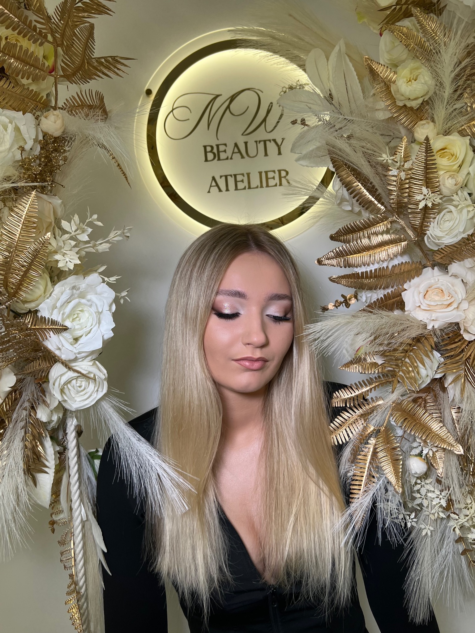 Portret kobiety z blond włosami i delikatnym, błyszczącym upiększeniem oczu, otoczonej dekoracją z białych i złotych kwiatów oraz piór na tle logo salonu 'MW Beauty Atelier'.