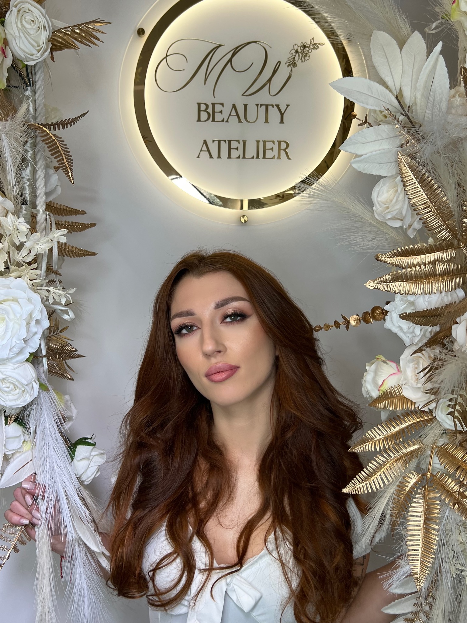 Portret kobiety z długimi, rudymi włosami i delikatnym upiększeniem, pozującej na tle dekoracji z białych i złotych kwiatów oraz logo salonu 'Beauty Atelier'.