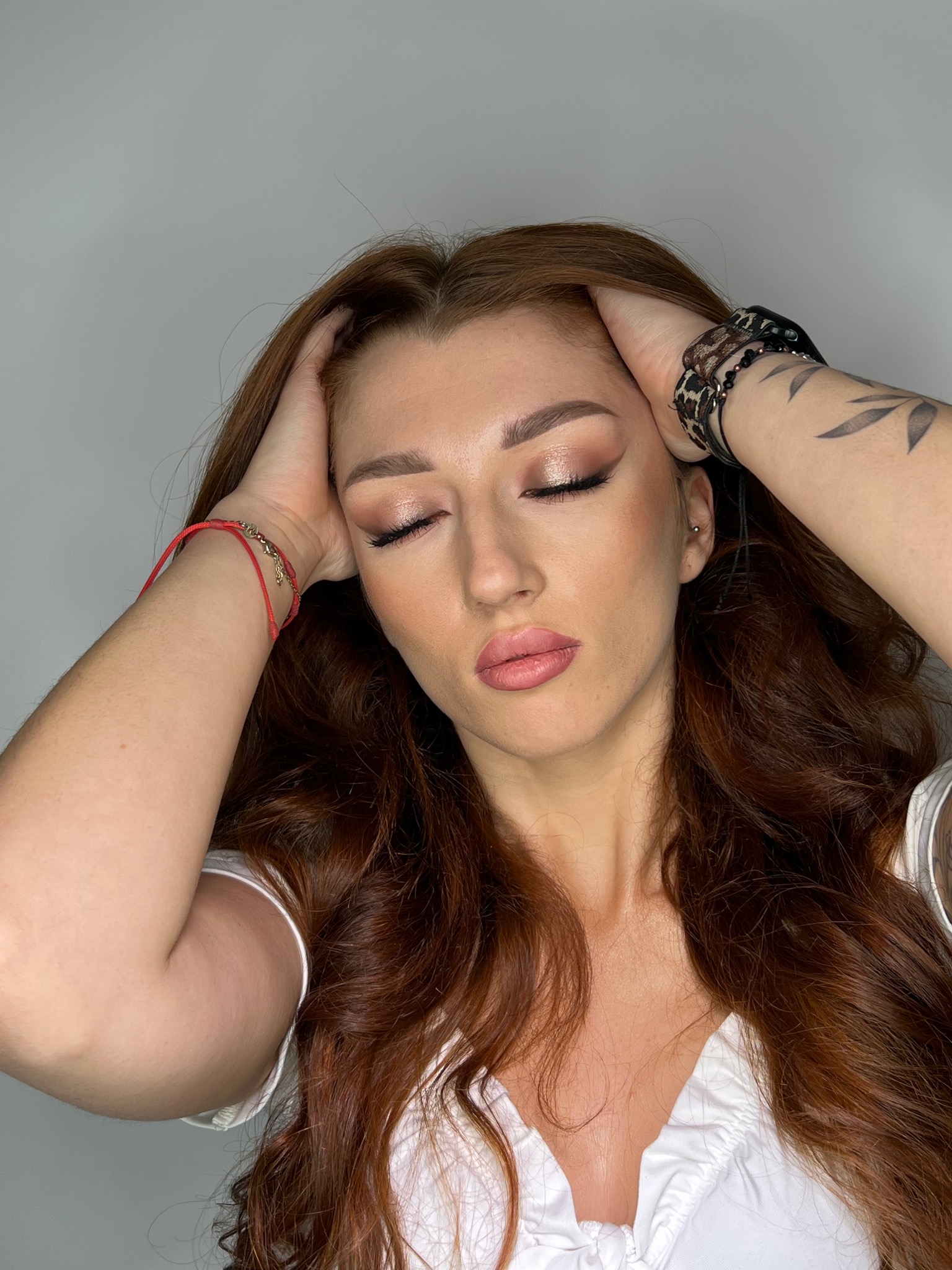 Portret kobiety z zamkniętymi oczami, delikatnym makijażem oczu i ust, dłońmi uniesionymi do głowy, widoczne tatuaże na ramieniu i czerwona bransoletka na nadgarstku.
