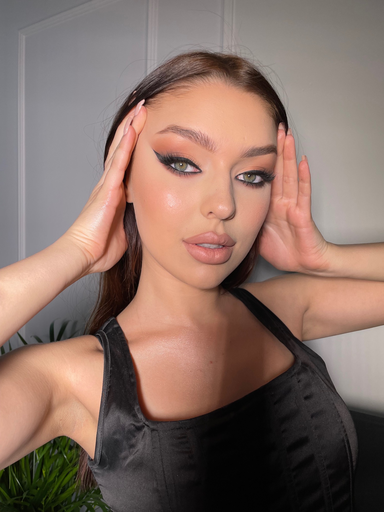 Portret kobiety z intensywnym makijażem oczu, w tym czarną kreską i brązowymi cieniami, z dłońmi uniesionymi do twarzy, eksponujący zielone oczy i satynową bluzkę.