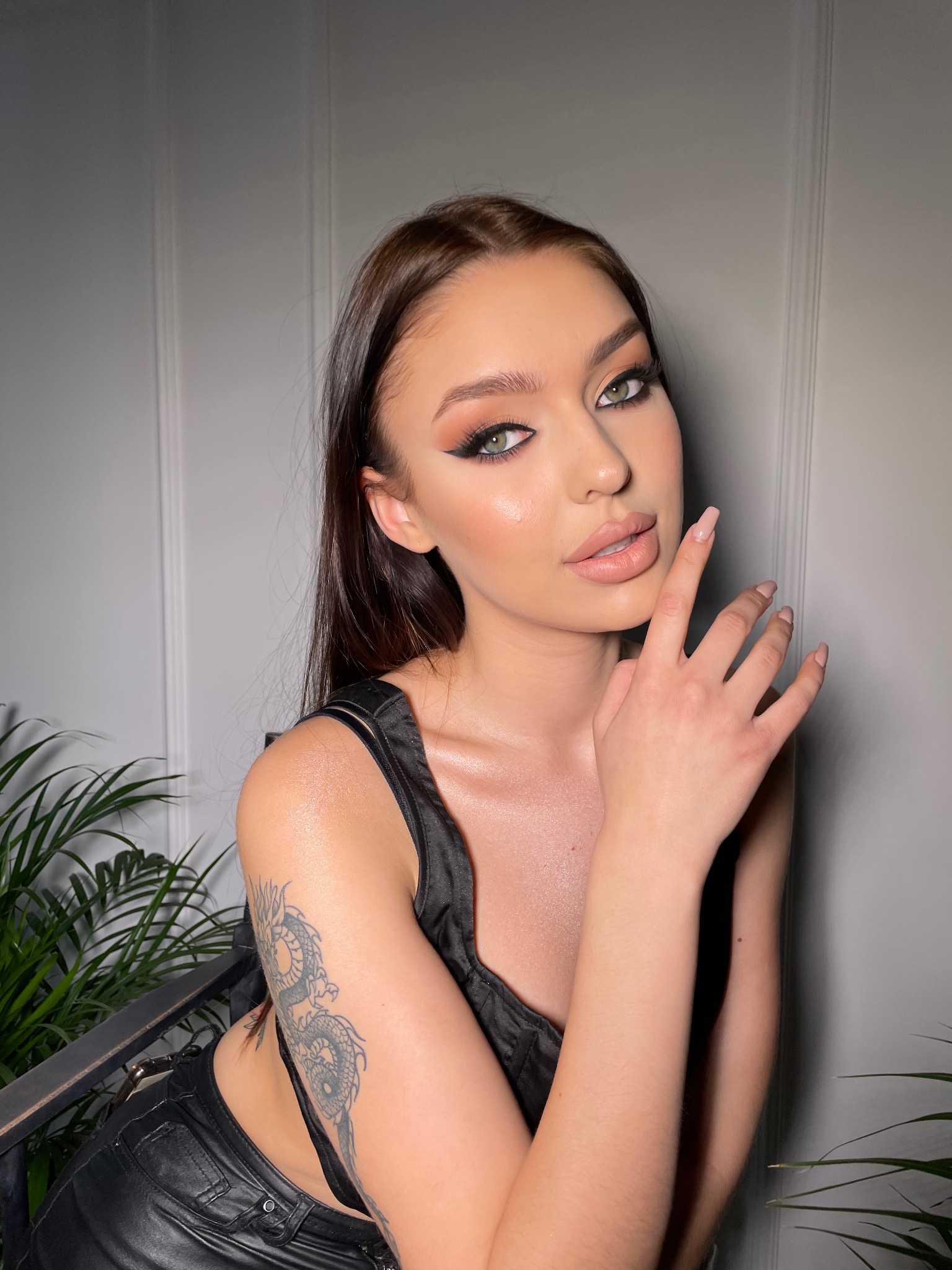 Portret kobiety z wyrazistym makijażem oczu i ust w odcieniach nude, dłoń przy twarzy, widoczny tatuaż smoka na ramieniu, tło stanowią rośliny doniczkowe i jasna ściana.