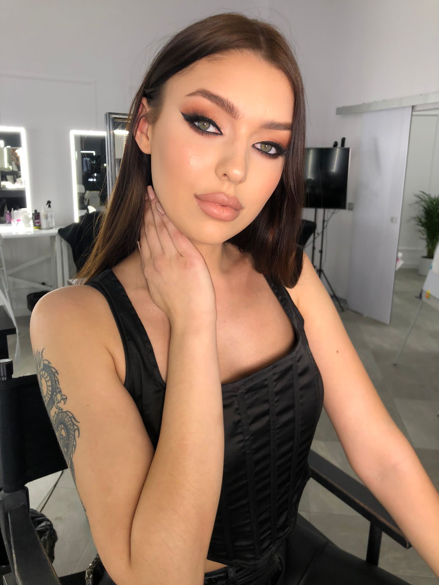 Portret kobiety z profesjonalnie wykonanym, wyrazistym makijażem oczu w odcieniach brązu i czerni, z widocznym tatuażem smoka na ramieniu, pozującej w studio.