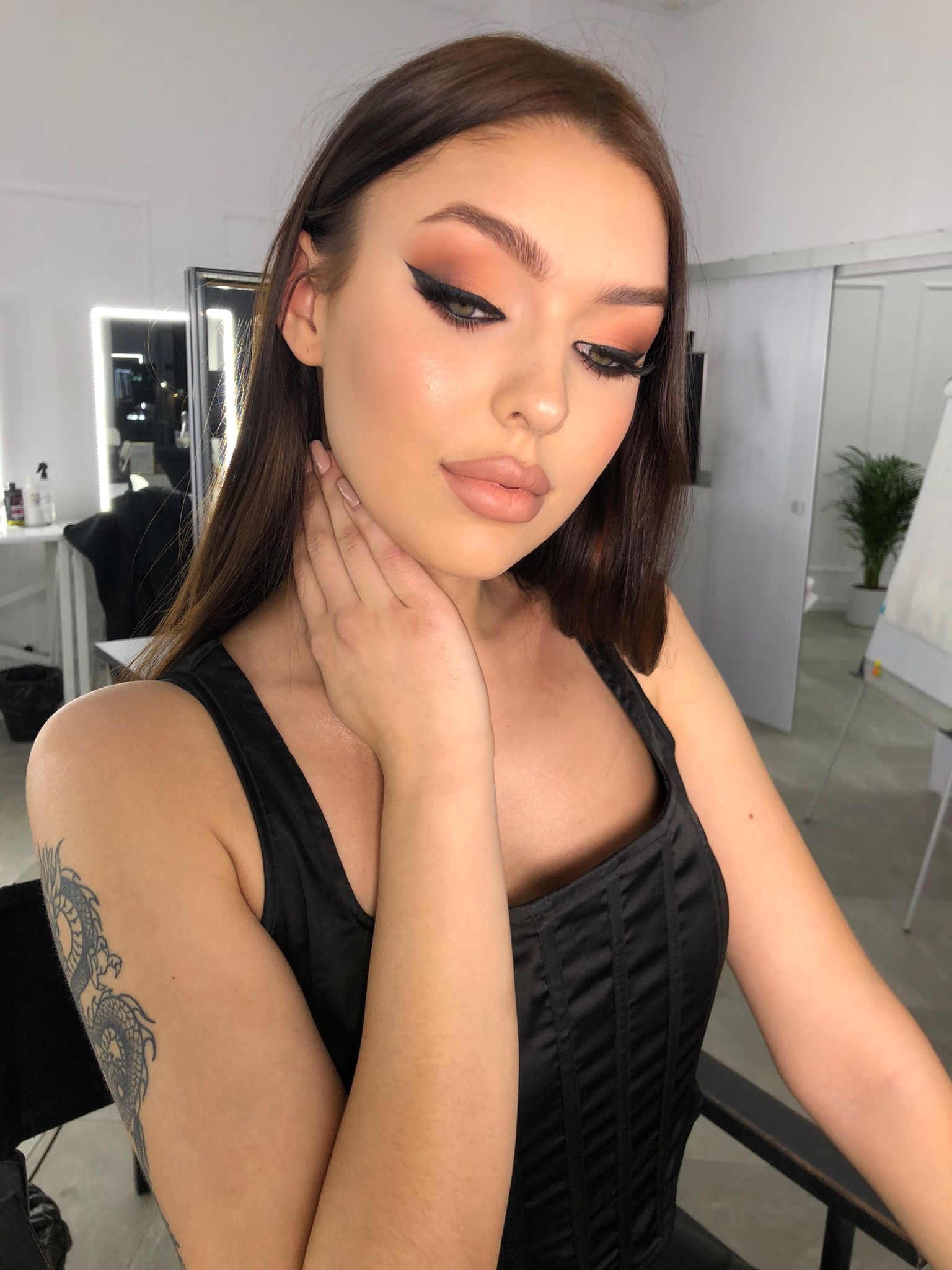 Portret kobiety z precyzyjnym, czarnym eyelinerem i brzoskwiniowymi cieniami na powiekach, w czarnym gorsecie, z tatuażem smoka na ramieniu, pozującej w salonie kosmetycznym.