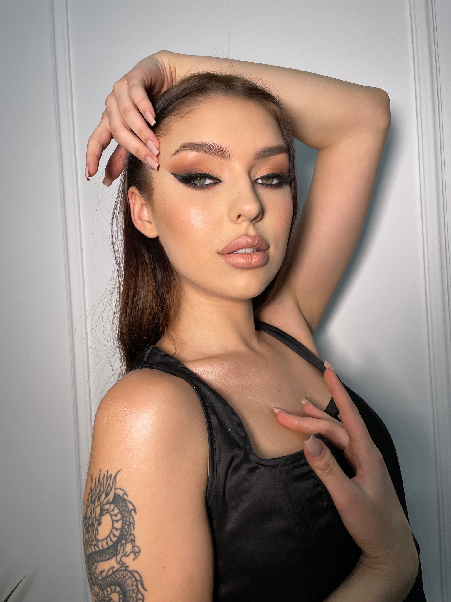 Portret kobiety z profesjonalnie wykonanym makijażem oczu, brwi i ust, z tatuażem smoka na ramieniu, pozującej na tle białych drzwi.