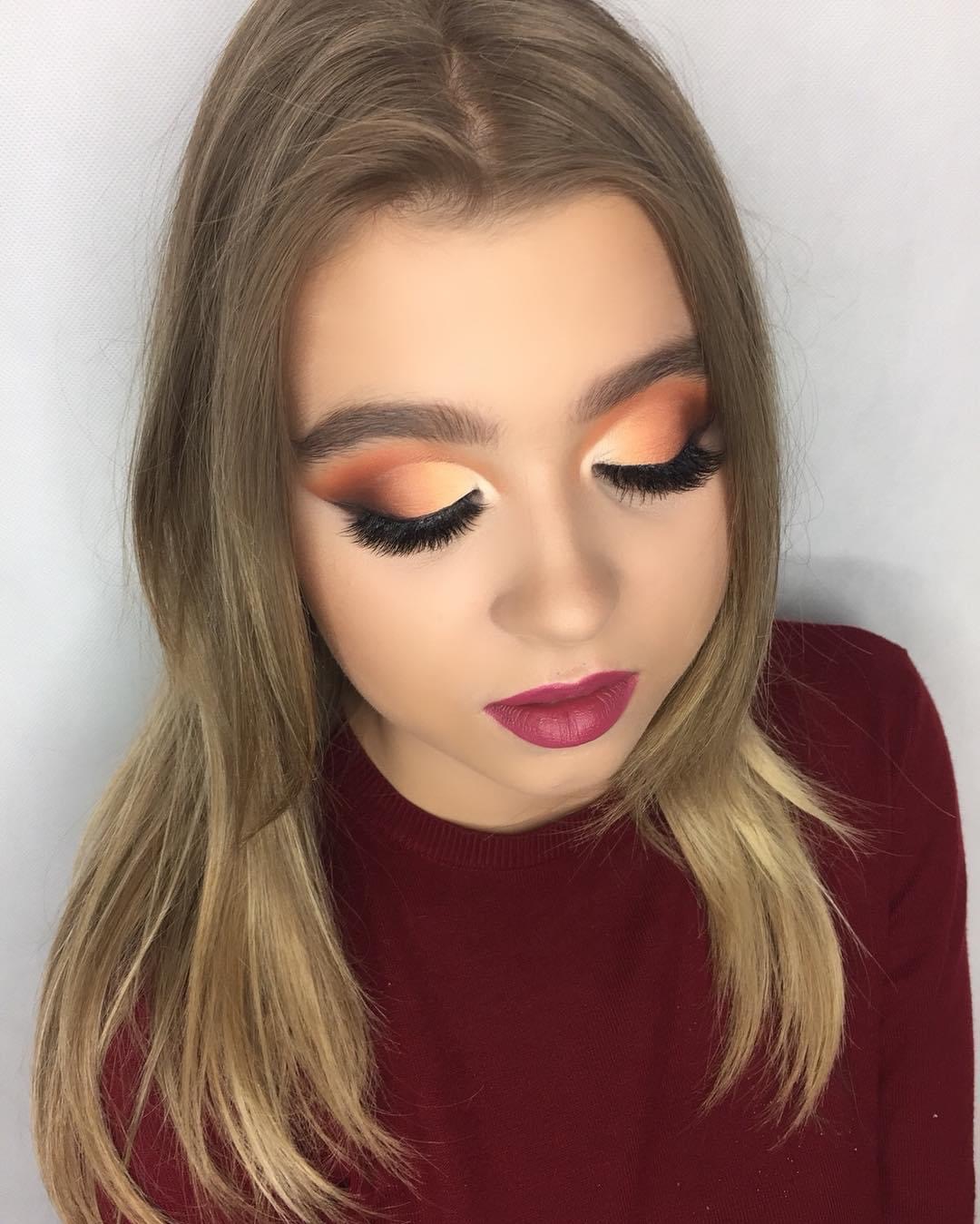 Portret kobiety z zamkniętymi oczami prezentującej makijaż oczu w odcieniach pomarańczy i brązu z wyraźnie podkreślonymi, sztucznymi rzęsami oraz bordową pomadką na ustach, na białym tle.