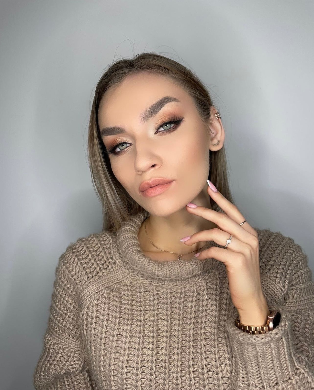 Portret kobiety z profesjonalnym make-upem oczu w odcieniach brązu i beżu, delikatny róż na policzkach i ustach, dłonie z biżuterią przy twarzy, sweter z golfem.