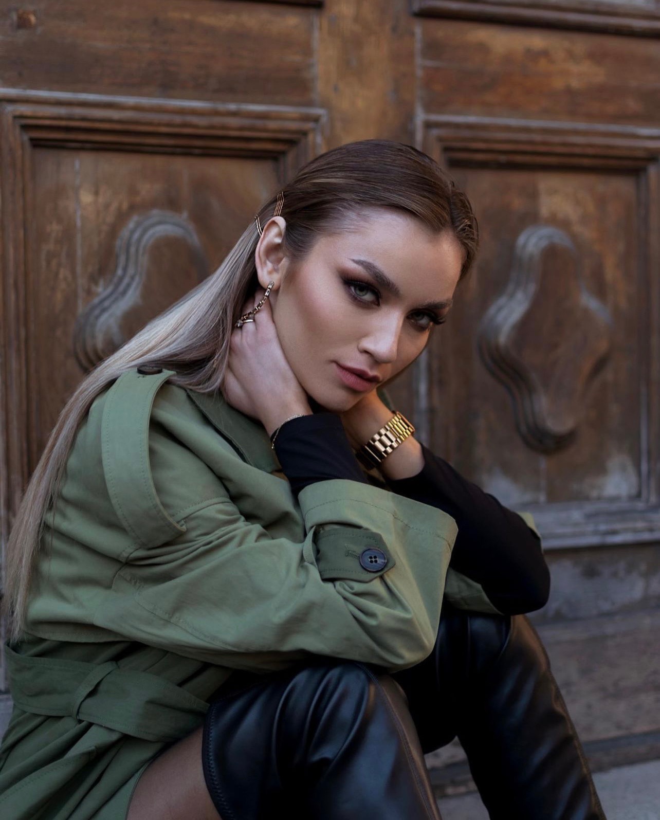 Portret kobiety w Jeleniej Górze z profesjonalnie wykonanym makijażem oczu, podkreślonymi brwiami, subtelnymi ustami, złotymi spinkami we włosach i złotym zegarkiem, pozującej na tle drewnianych...