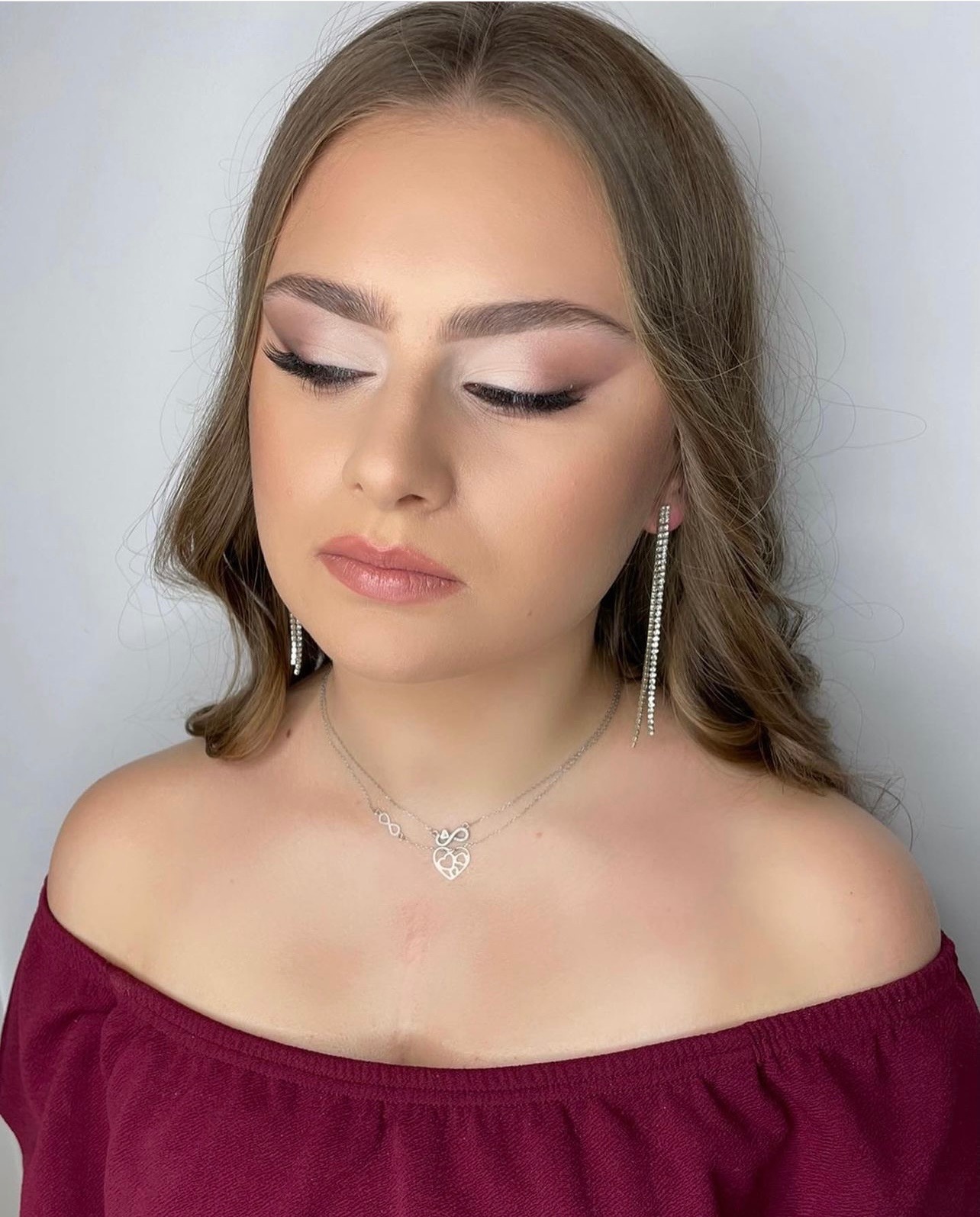 Portret młodej kobiety z zamkniętymi oczami, prezentującej delikatny, dzienny w odcieniach beżu i brązu, z błyszczącymi kolczykami i naszyjnikiem z zawieszką w kształcie serca.