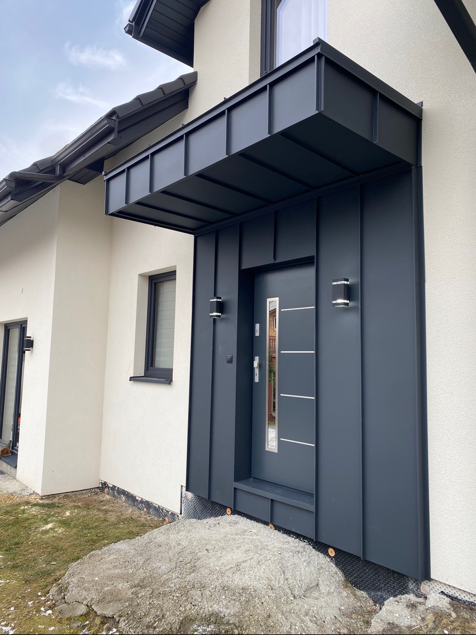 Nowoczesne wejście do domu z ciemnoszarą blachą trapezową, osłonięte zadaszeniem z tego samego materiału, oświetlone dwoma kinkietami, z widoczną rynną i fragmentem dachu. Na pierwszym planie teren...
