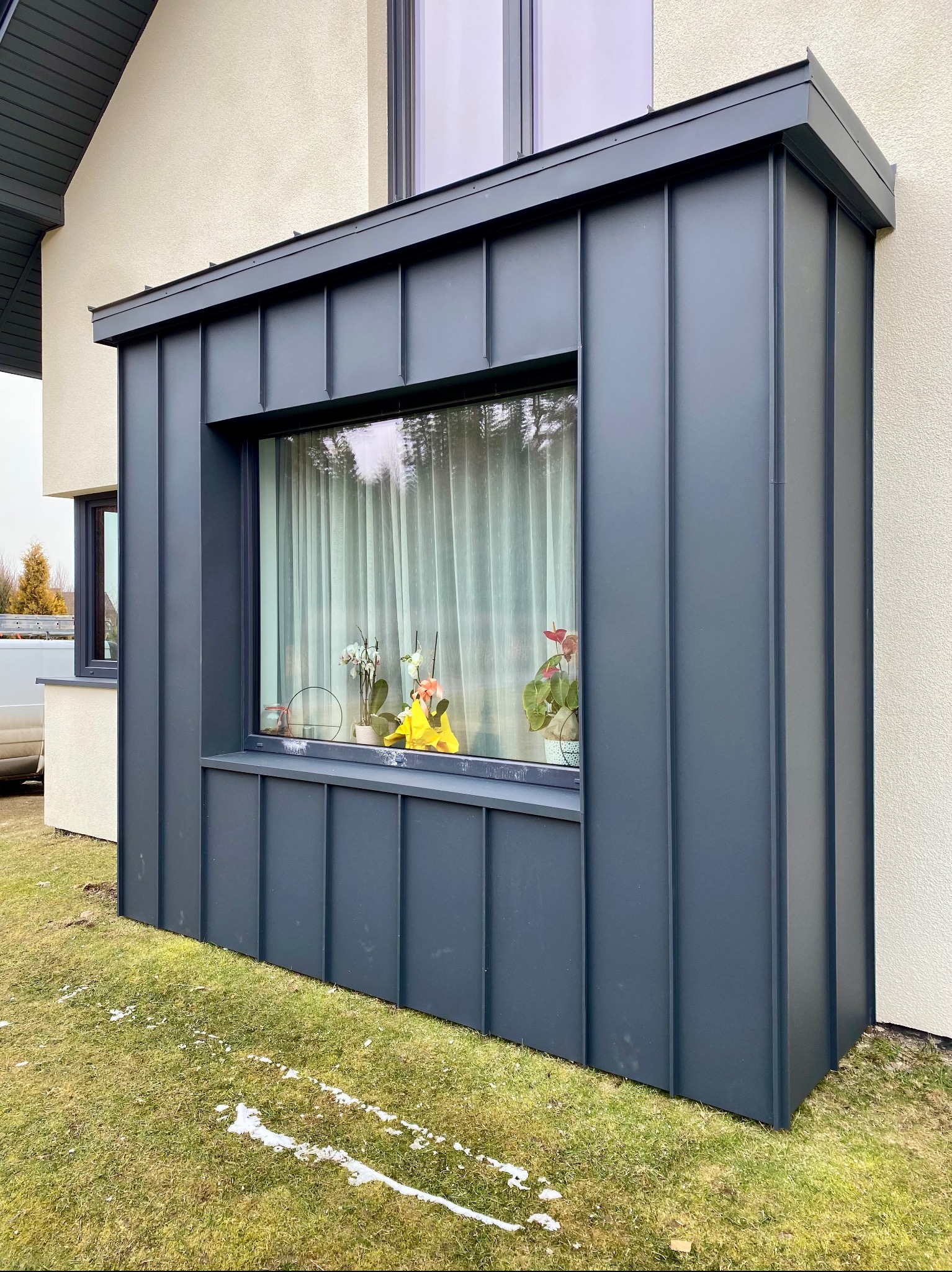 Nowoczesna dobudówka do domu z ciemnoszarej blachy trapezowej, widok z zewnątrz, z oknem i roślinami na parapecie.