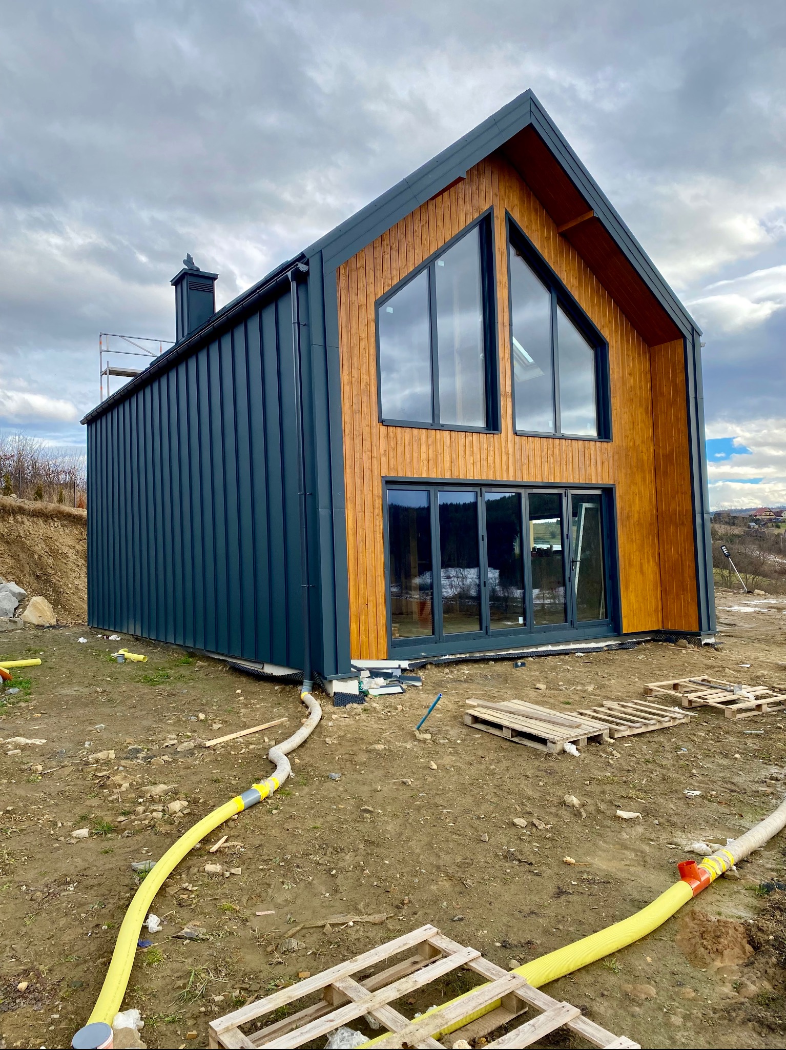 Nowoczesny dom z drewnianą elewacją i ciemnymi panelami, widoczne duże okna i przesuwne drzwi, teren wokół w trakcie prac wykończeniowych, żółte rury na ziemi, palety budowlane.