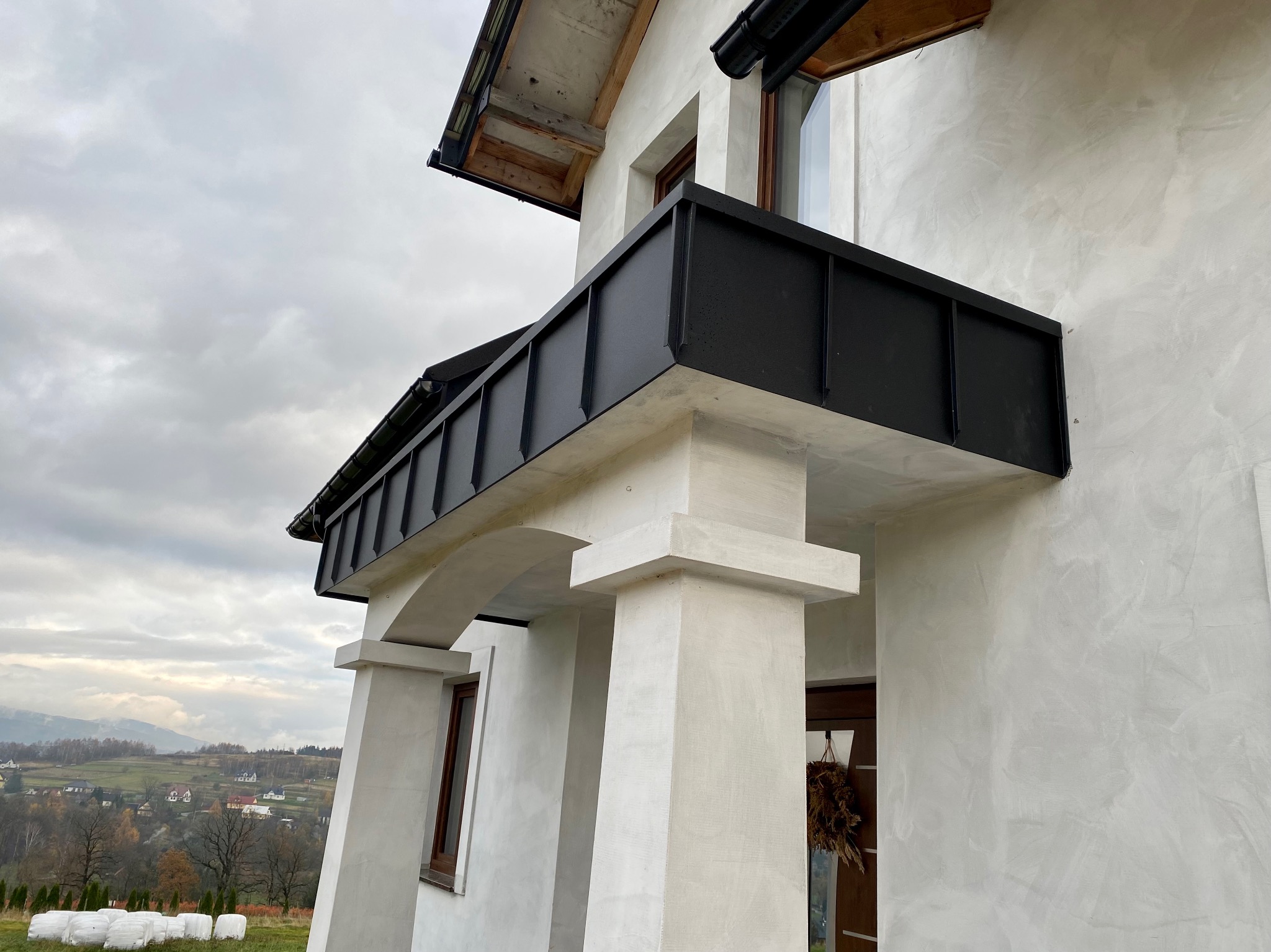 Nowoczesny dom z balkonem wykończonym czarną blachą, widok z dołu na elewację w jasnym odcieniu i elementy konstrukcyjne, w tle pagórkowaty krajobraz.