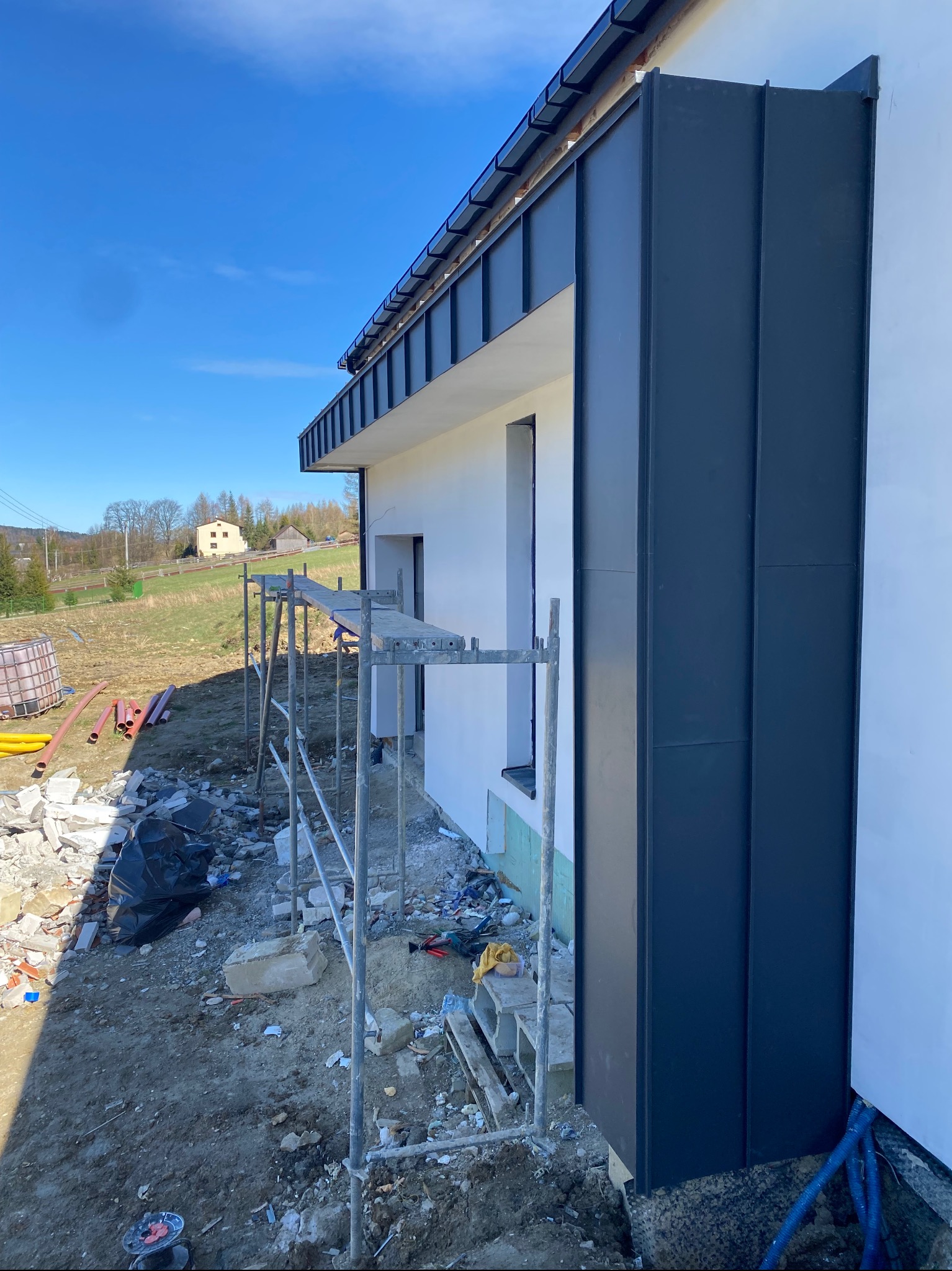 Nowoczesny dom z elewacją z ciemnej blachy na etapie wykończenia, widoczne rusztowanie, niezagospodarowany teren wokół budynku i błękitne niebo.