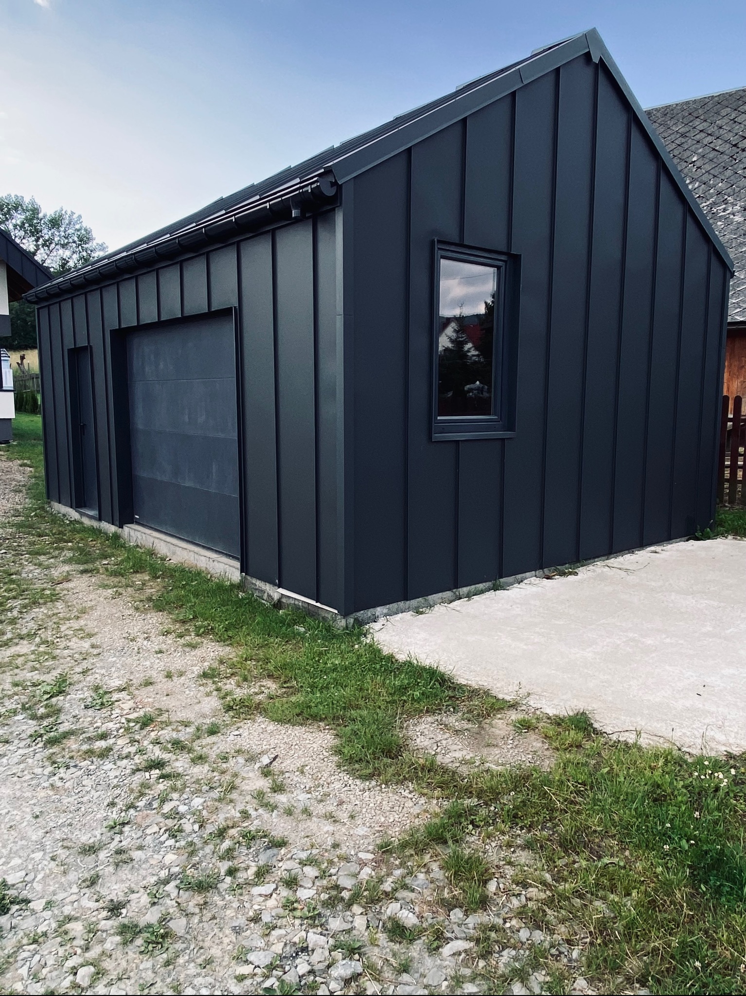 Nowoczesny, ciemnoszary budynek gospodarczy z metalową elewacją, widoczne rynny, okno i brama garażowa, otoczenie z kamienistą ścieżką i trawą.