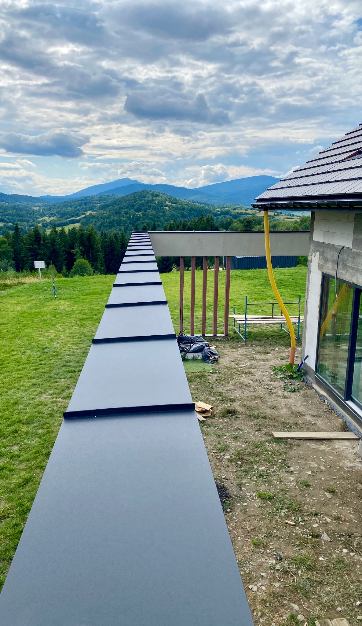 Nowoczesny dom z ciemną blachą na dachu, widok z góry na tle górskiego krajobrazu. Żółta rynna odprowadza wodę. Widoczne elementy konstrukcyjne.