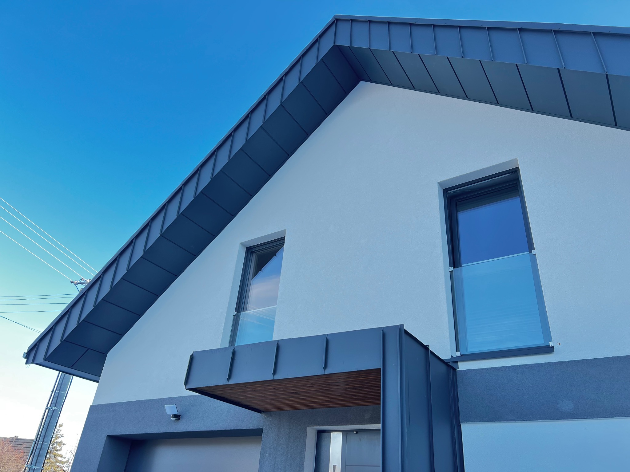 Nowoczesny dom z blachy trapezowej w kolorze grafitowym, z widocznym fragmentem elewacji i oknami. Ujęcie z dołu, podkreślające geometrię dachu i minimalistyczny design.