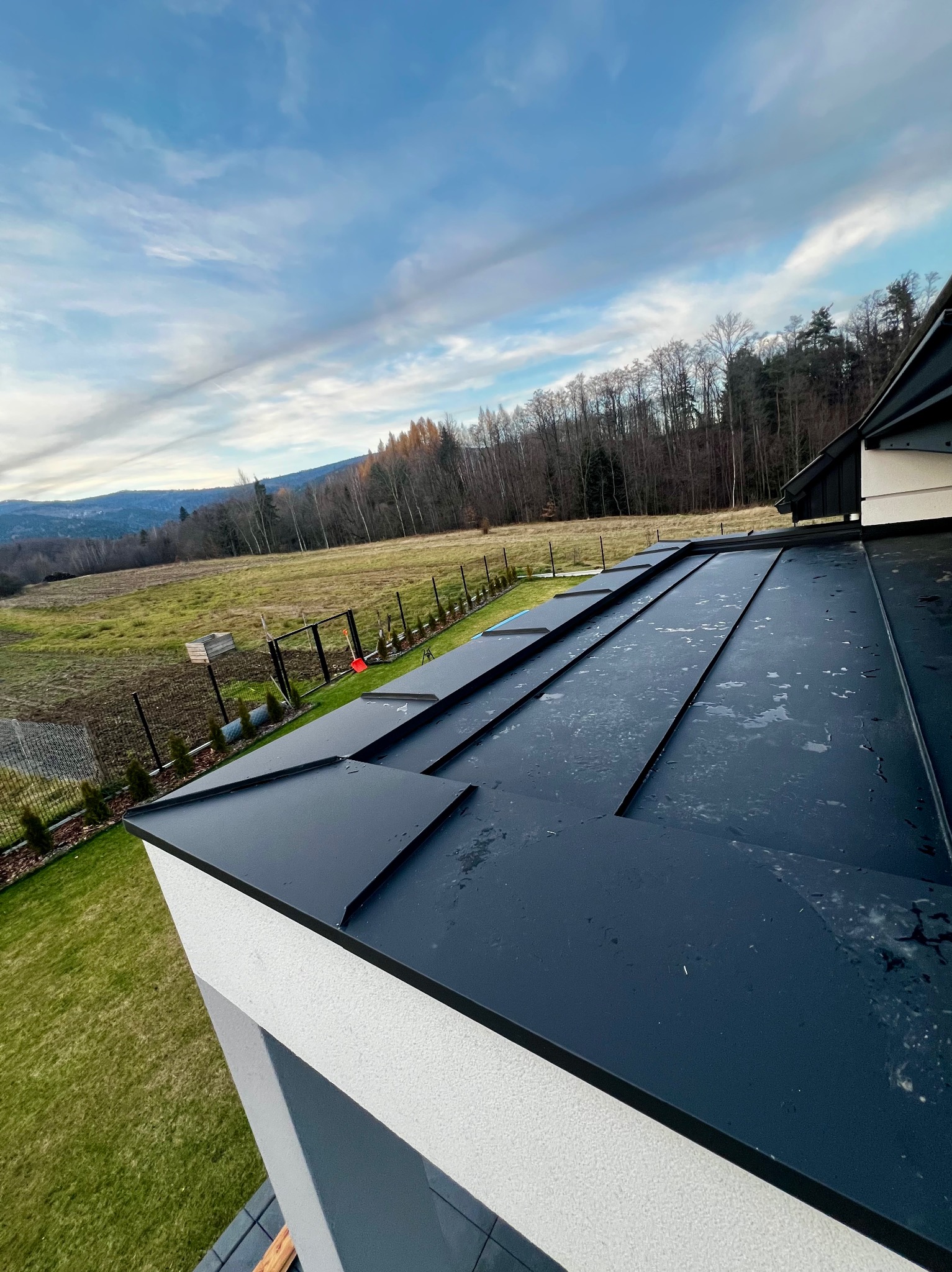 Czarny, metalowy dach z widokiem na zielony krajobraz i błękitne niebo z chmurami. Widoczne elementy wykończenia dachu i fragment elewacji budynku.