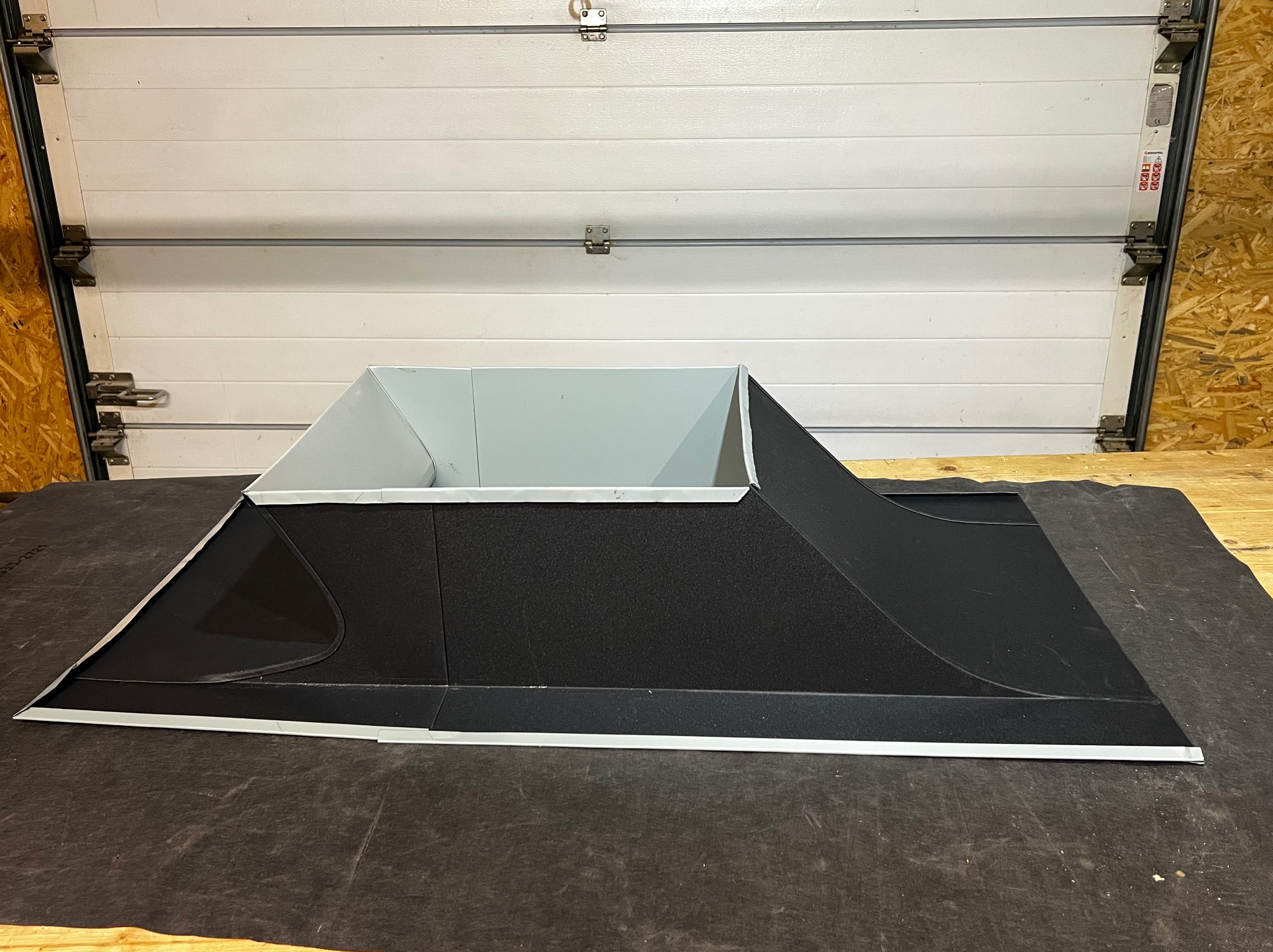 Model fragmentu dachu z obróbką blacharską w warsztacie, ukazujący detale konstrukcyjne i materiałowe. Widoczna blacha w kolorze czarnym i szarym. Tło stanowi brama garażowa.